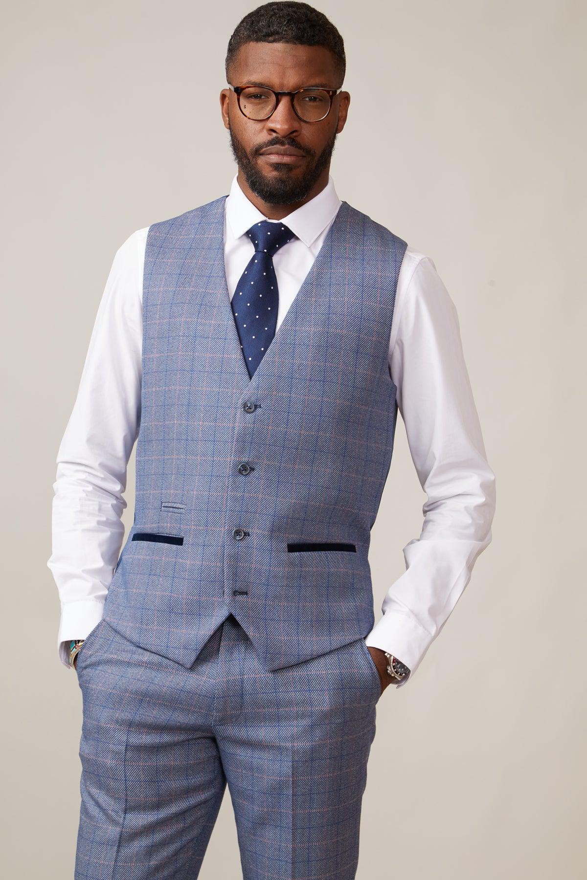 A Black man with glasses and a beard wears a blue plaid suit, white shirt, navy polka-dot tie, and the marcdarcy HILTON Blue Tweed Check Single Breasted Waistcoat. His hands rest in his pockets.