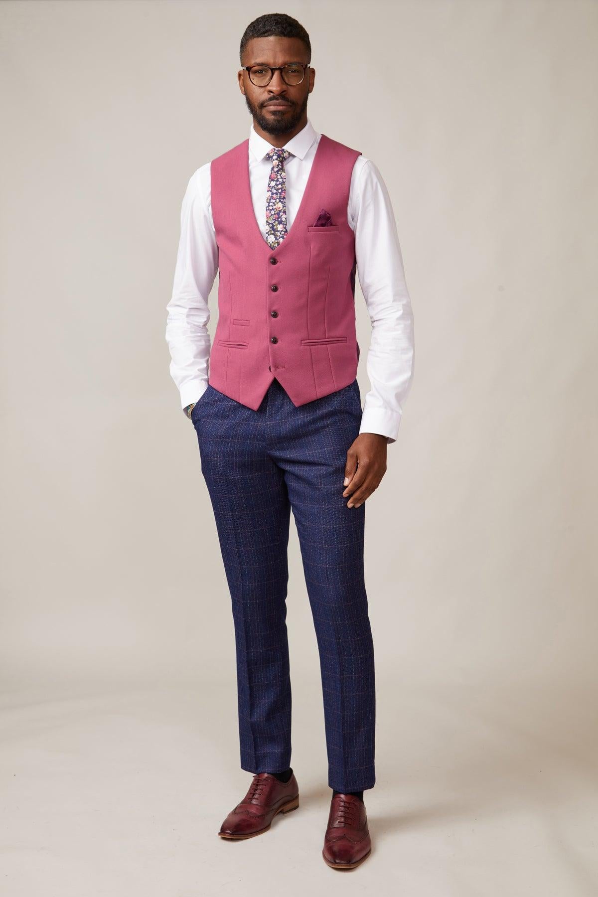 A man with a beard and glasses stands against a plain background wearing the marcdarcy KELVIN Berry Single Breasted Waistcoat, white shirt, floral tie, blue plaid trousers, and burgundy shoes—one hand in his pocket for a sharp summer wedding look.