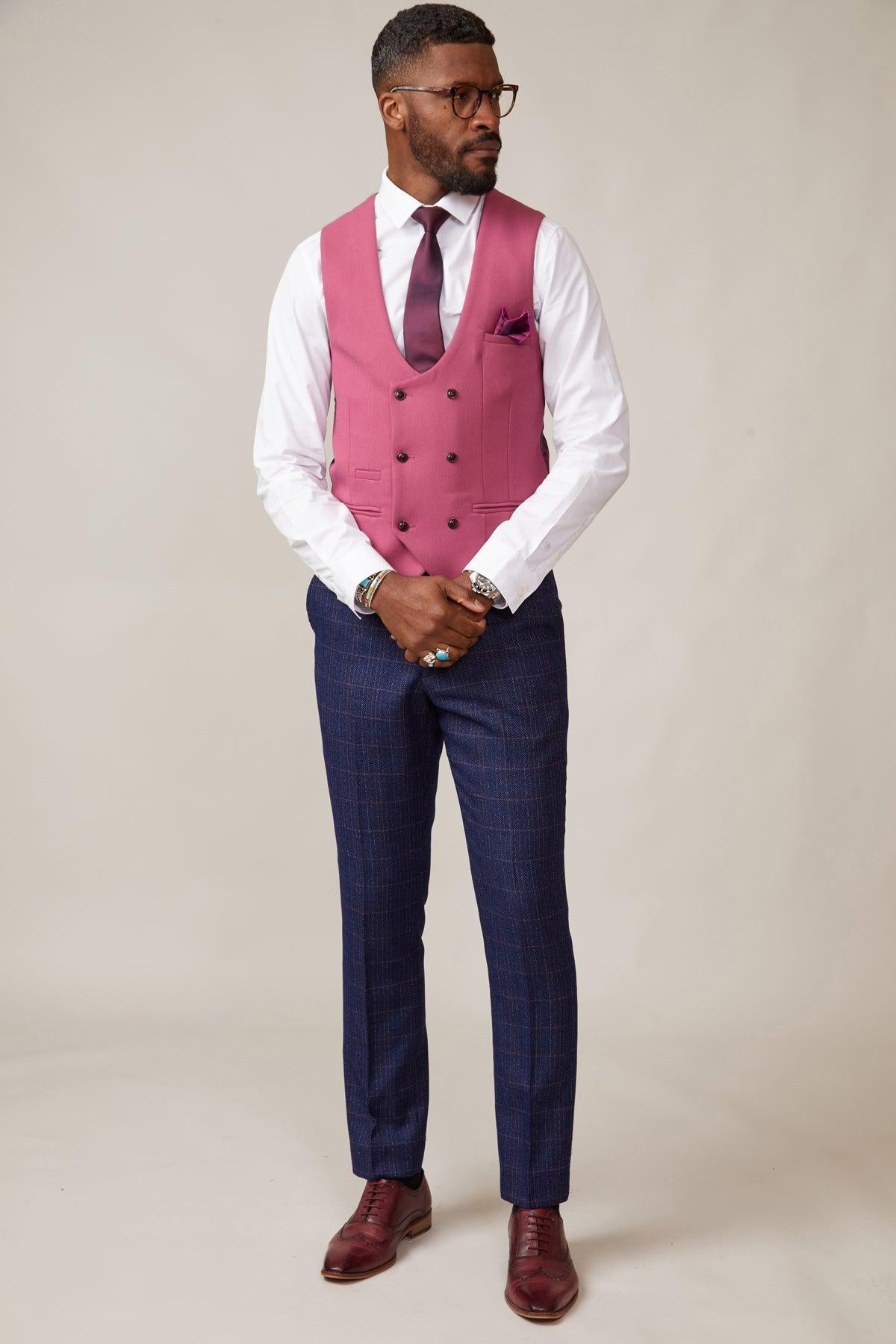 A man stands against a plain background wearing marcdarcy’s KELVIN - Berry Double Breasted Waistcoat with a white shirt, matching tie and pocket square, blue checked trousers, brown dress shoes, and glasses while looking to the side.