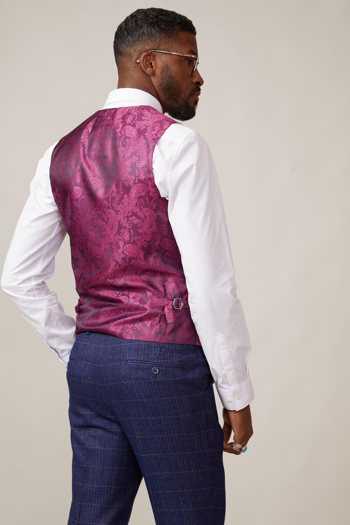 A man faces away from the camera, wearing a white shirt and marcdarcy’s KELVIN - Berry Double Breasted Waistcoat with a paisley pattern, paired with blue checkered dress pants. He also wears glasses, a ring, and has one hand in his pocket.