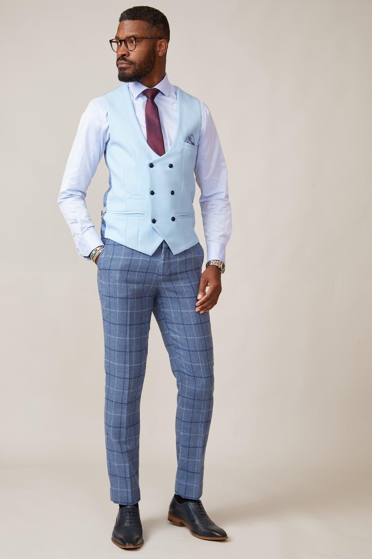 A man in the marcdarcy KELVIN Sky Blue Double Breasted Waistcoat, white shirt, burgundy tie, blue plaid trousers, and black dress shoes stands against a neutral background, looking to the side with one hand in his pocket.