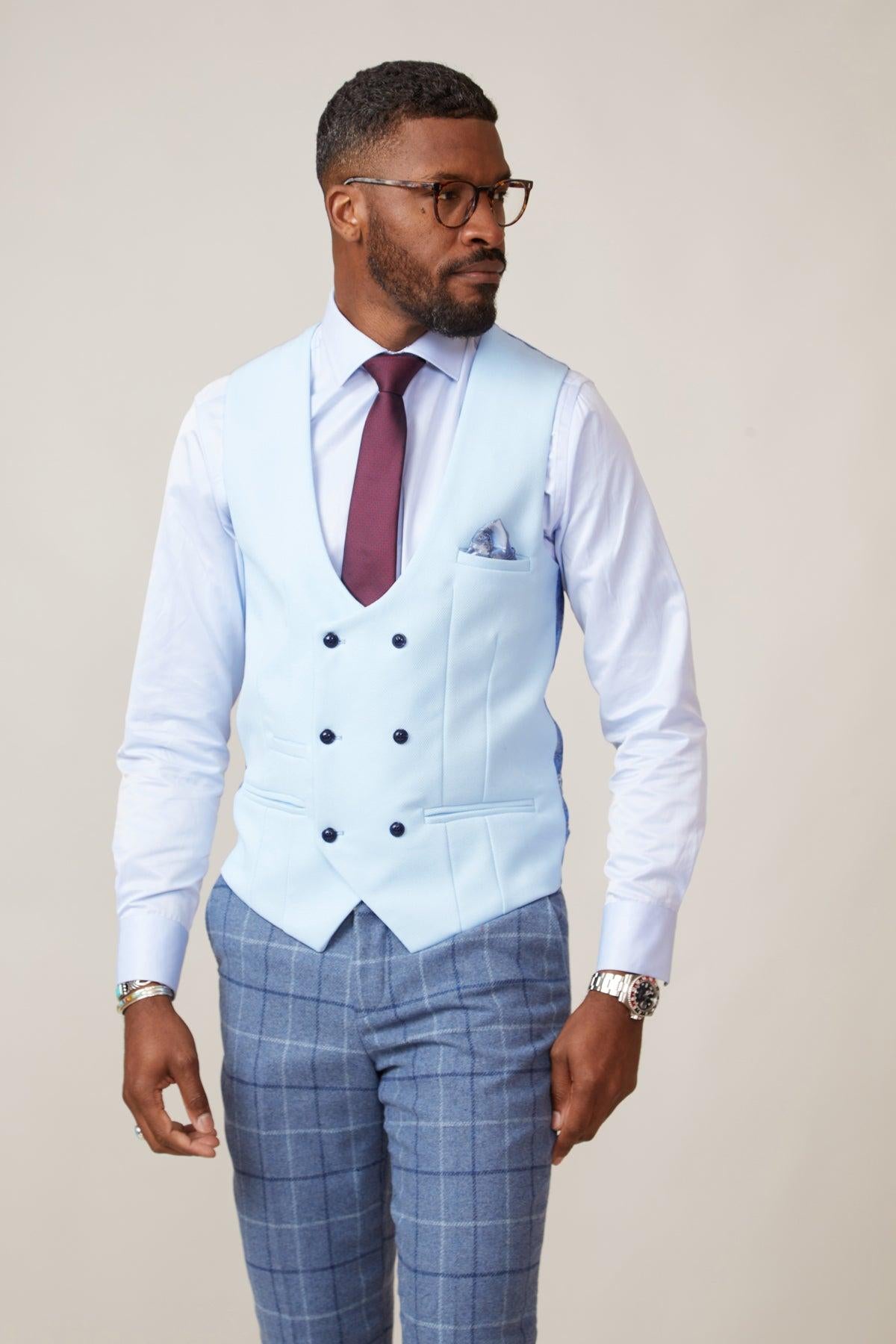 A man dressed in the marcdarcy KELVIN Sky Blue Double Breasted Waistcoat, white shirt, burgundy tie, blue plaid trousers, wristwatch, and glasses stands against a simple backdrop, looking thoughtfully to his left.
