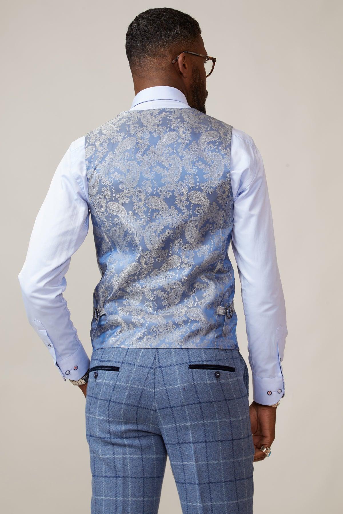A man with short hair and glasses is seen from behind wearing the marcdarcy KELVIN Sky Blue Double Breasted Waistcoat over a white dress shirt and light blue plaid trousers, his hands relaxed by his sides.