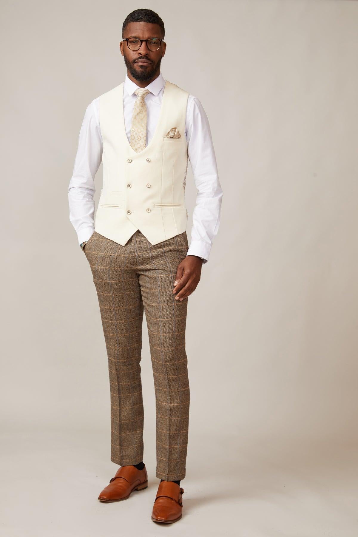 A man stands against a plain background wearing the marcdarcy KELVIN Cream Double Breasted Waistcoat, a white shirt, beige patterned tie, brown plaid pants, and brown dress shoes. He has short hair, a beard, glasses, and his hands in his pockets.
