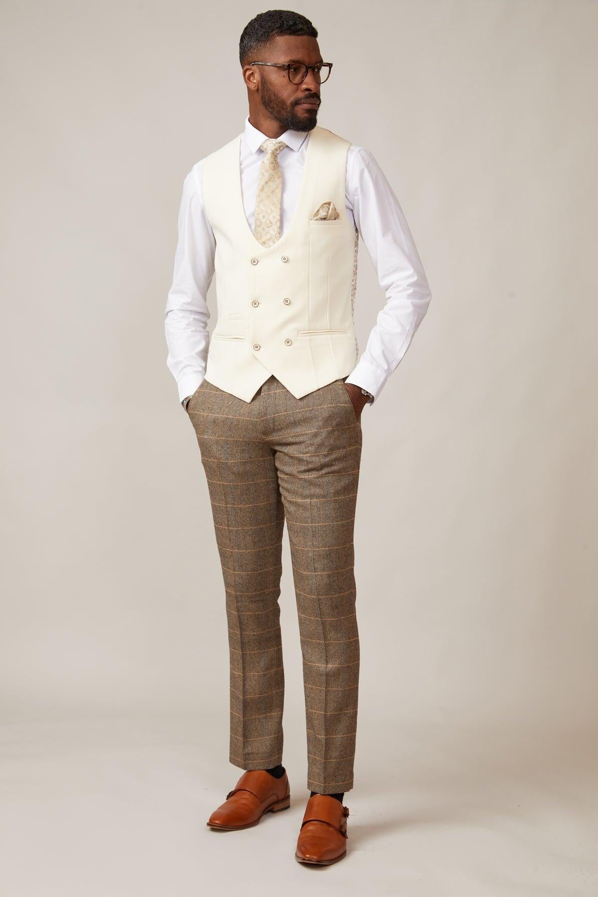 A man stands against a plain light background, hands in pockets, wearing the marcdarcy KELVIN cream double-breasted waistcoat with adjustable straps, a white dress shirt, beige patterned tie, and brown plaid trousers for a Traditional British look.