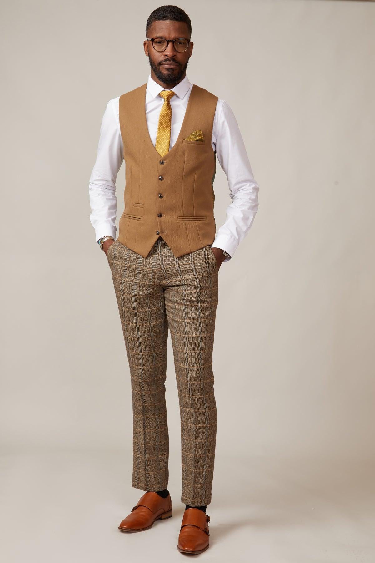 A man stands against a plain background, confidently showcasing the marcdarcy KELVIN - Oak Single Breasted Waistcoat over a white shirt with glasses, a yellow patterned tie, plaid pants, and brown dress shoes, hands in his pockets.
