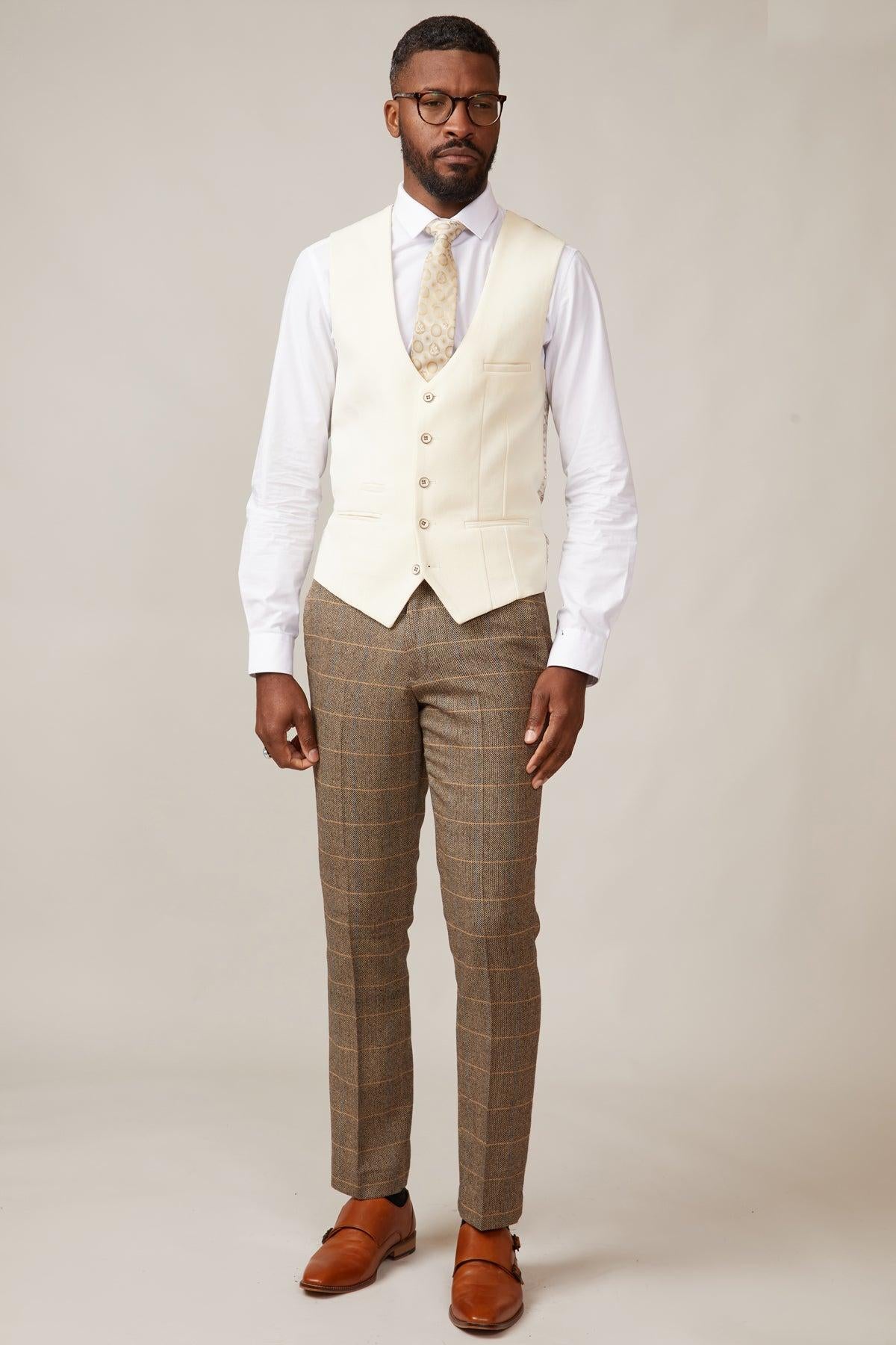 A man wears the marcdarcy KELVIN cream single breasted waistcoat over a white shirt with a patterned tie, brown plaid trousers, and brown double monk strap shoes. He has short hair, a beard, glasses, and stands against a plain background.