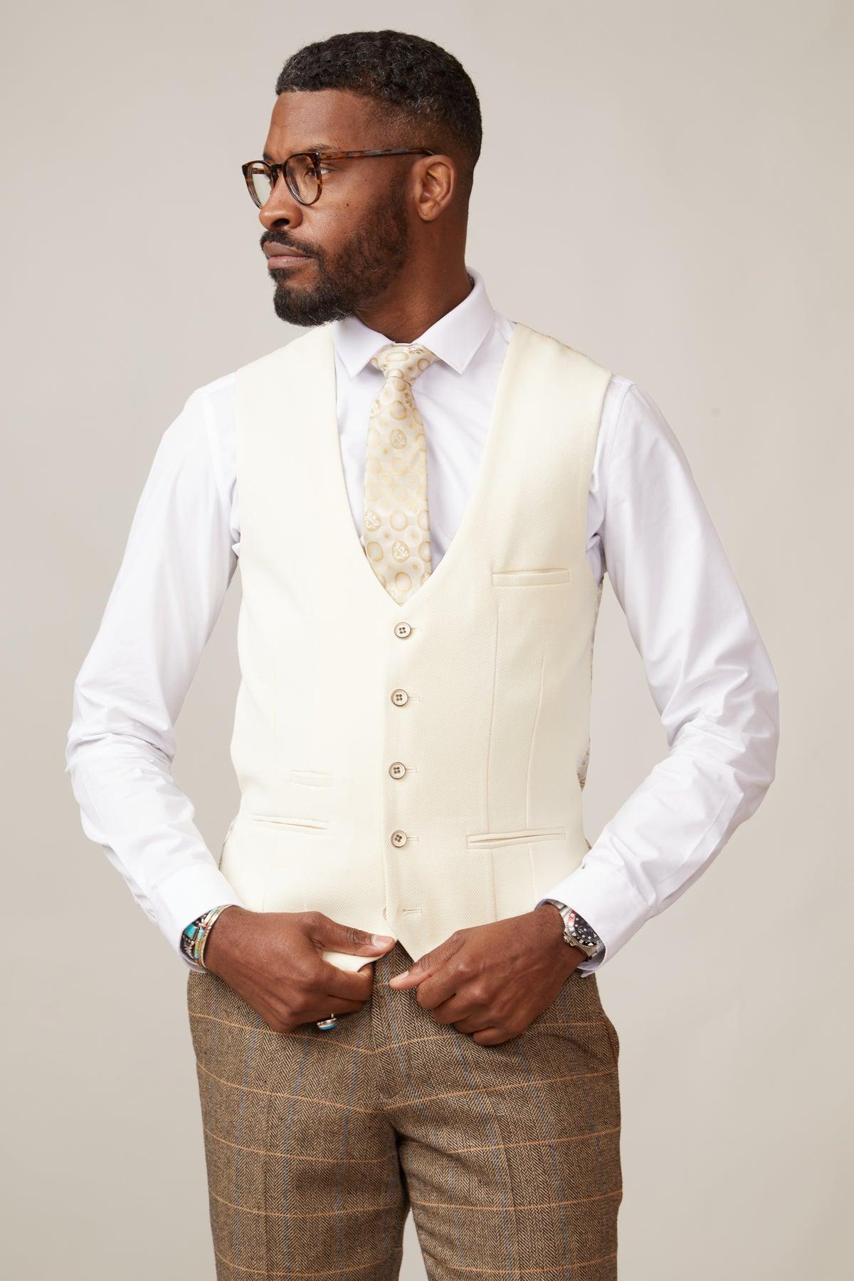 A man with short hair, a beard, and glasses wears a slim fit white dress shirt, marcdarcy KELVIN - Cream Single Breasted Waistcoat, patterned tie, and brown plaid pants inspired by British tailoring, standing turned slightly to the side.