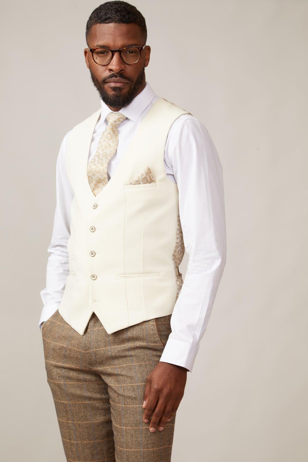 A man wearing glasses, a white shirt, marcdarcy's KELVIN cream single breasted waistcoat with matching tie and pocket square, and brown plaid trousers stands against a light background.