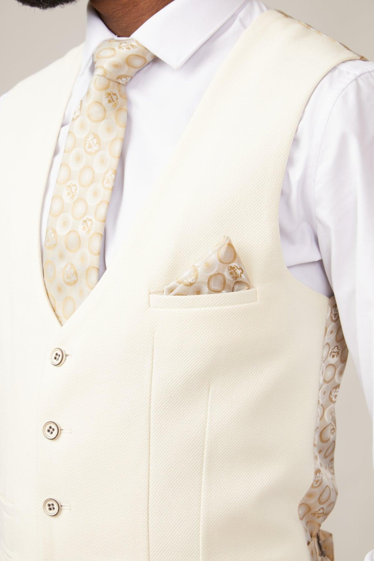 A man wears the marcdarcy KELVIN Cream Single Breasted Waistcoat in a slim fit three-piece suit, paired with a matching patterned tie, pocket square, and white shirt. Textured fabric and light buttons evoke classic British style. His face is partly out of frame.