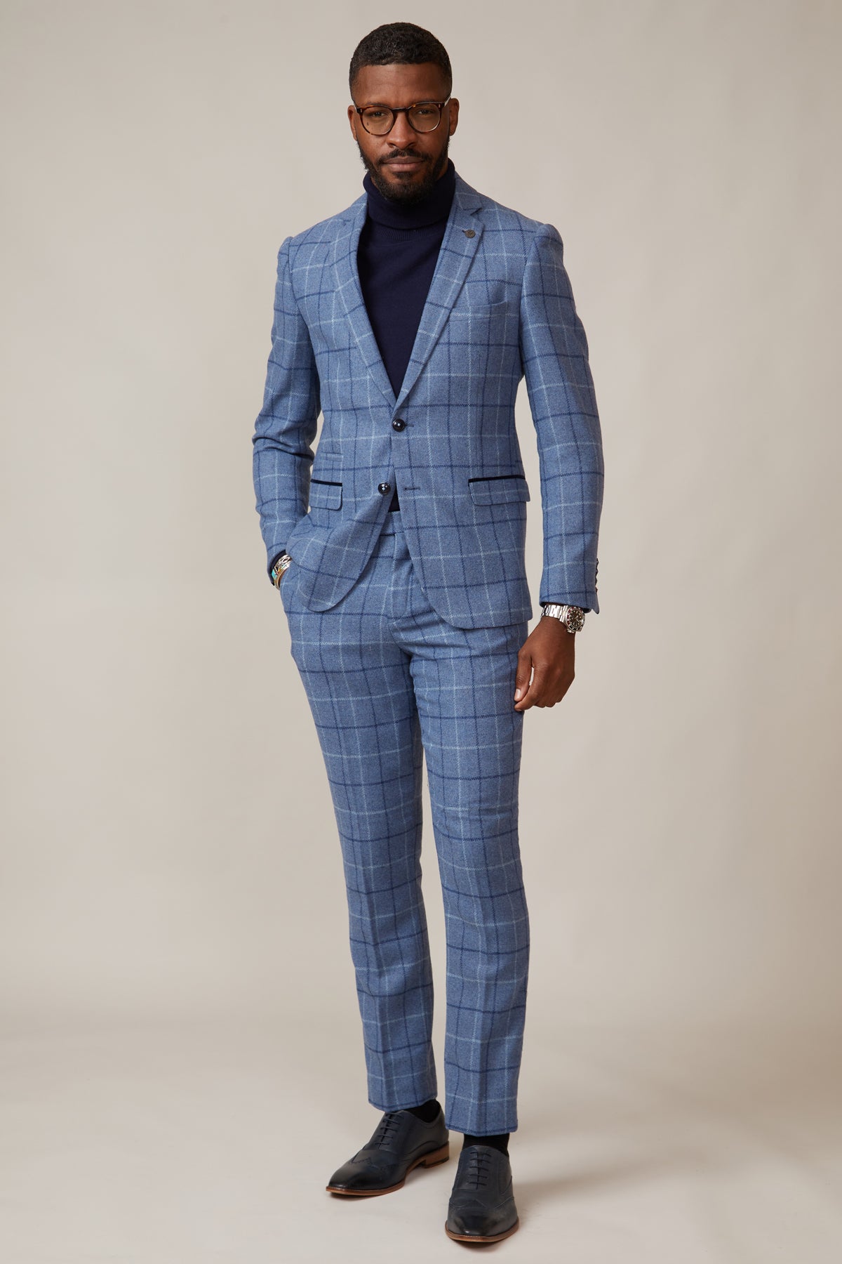 A Black man wearing the marcdarcy CLINTON Blue Tweed Check Blazer, a navy turtleneck, glasses, and black dress shoes.