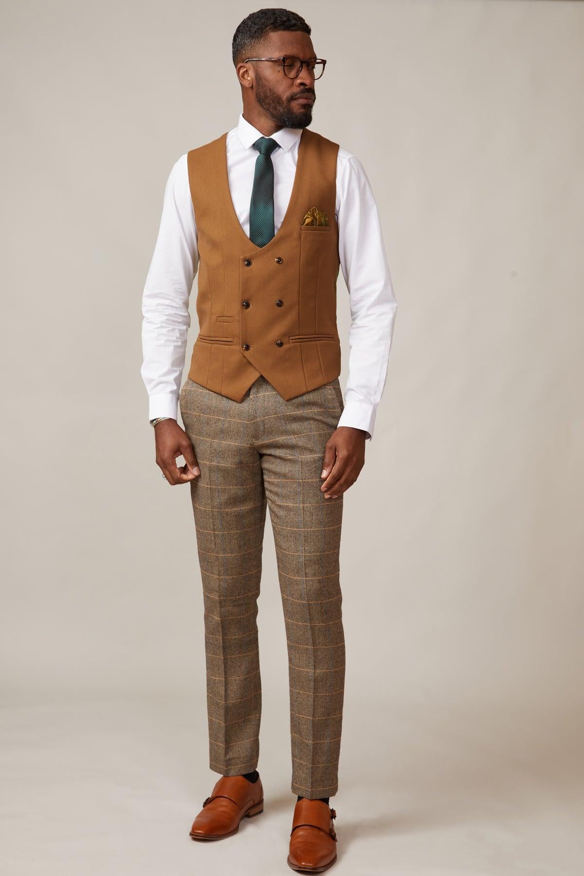 A Black man wears the marcdarcy KELVIN Tan Double Breasted Waistcoat, paired with a white shirt, green tie, plaid pants, and brown shoes, standing against a plain background.
