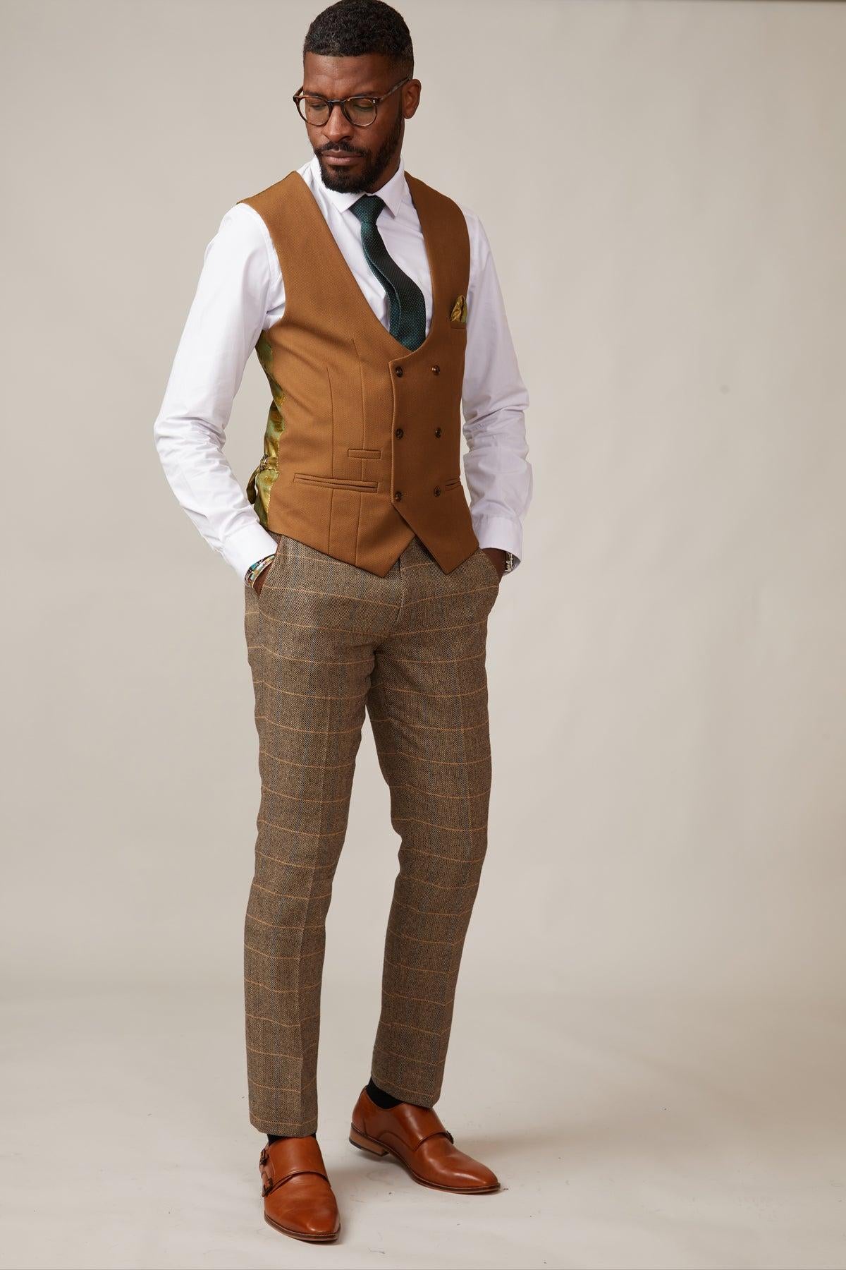 A man stands against a plain background wearing glasses, a white shirt, patterned trousers, a green tie, brown dress shoes, and the marcdarcy KELVIN Tan Double Breasted Waistcoat. His hands rest in his pockets as he looks downward.