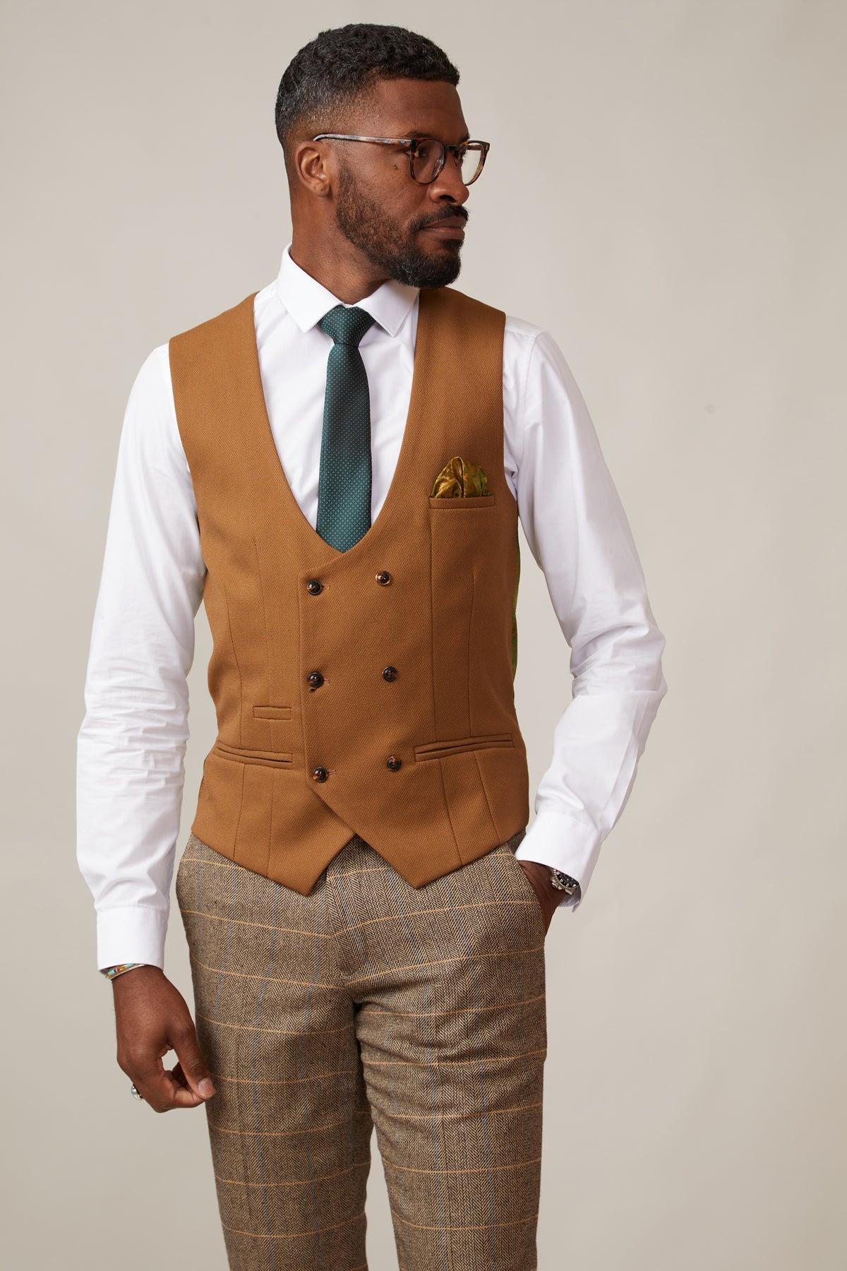 A man in glasses, a white shirt, the marcdarcy KELVIN Tan Double Breasted Waistcoat, a green tie, yellow patterned pocket square, and brown checked trousers stands against a plain background, looking to the side.