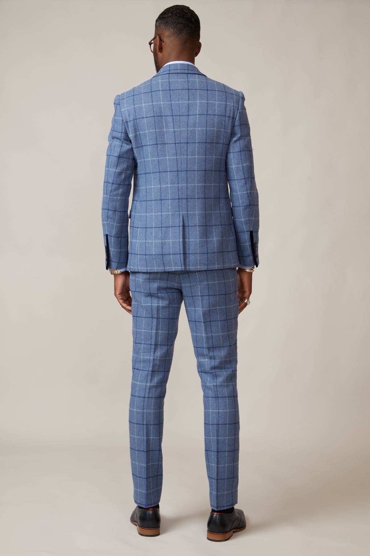 A man stands facing away from the camera in the marcdarcy CLINTON Blue Tweed Check Blazer and matching trousers, pairing dark shoes for a modern heritage look against a plain beige background.