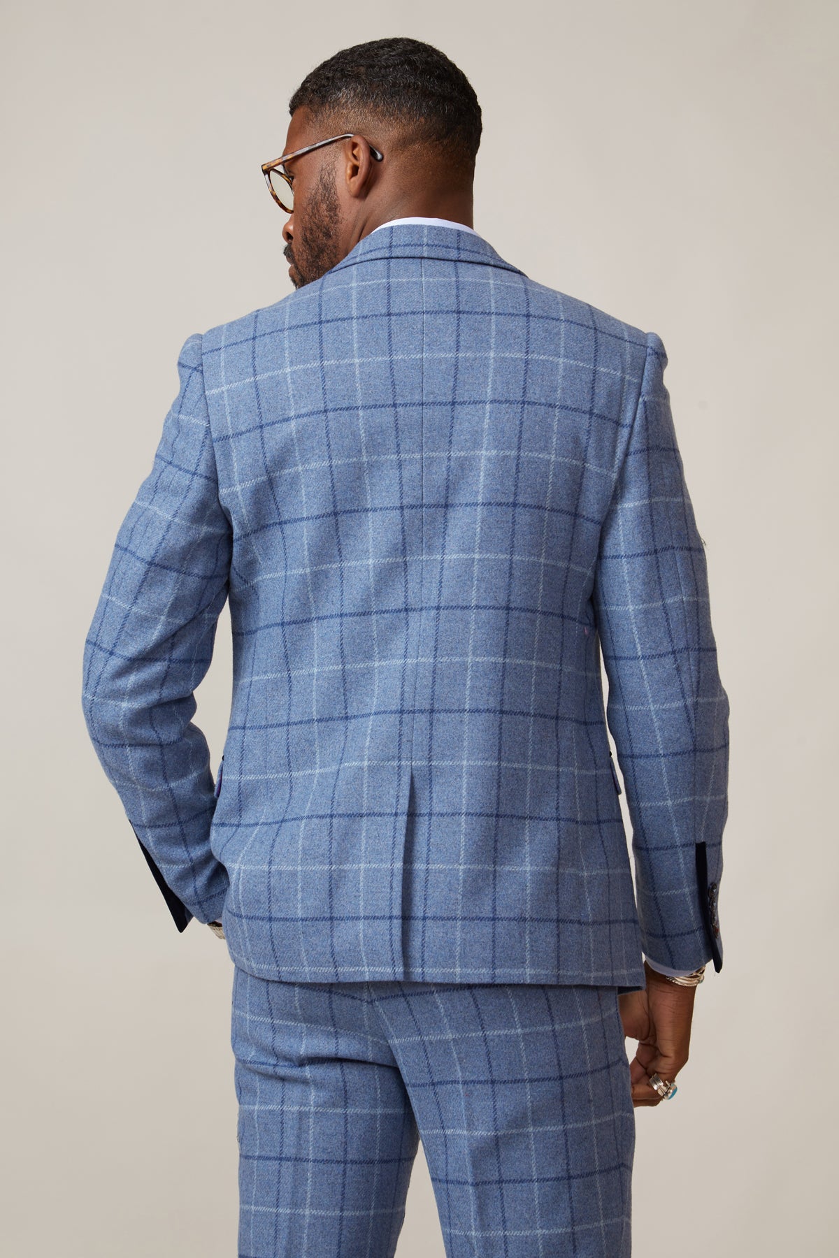 A man in a marcdarcy CLINTON Blue Tweed Check Blazer stands with his back to the camera, showcasing short hair, glasses, and a neat beard. His right hand rests by his side against a plain light background.