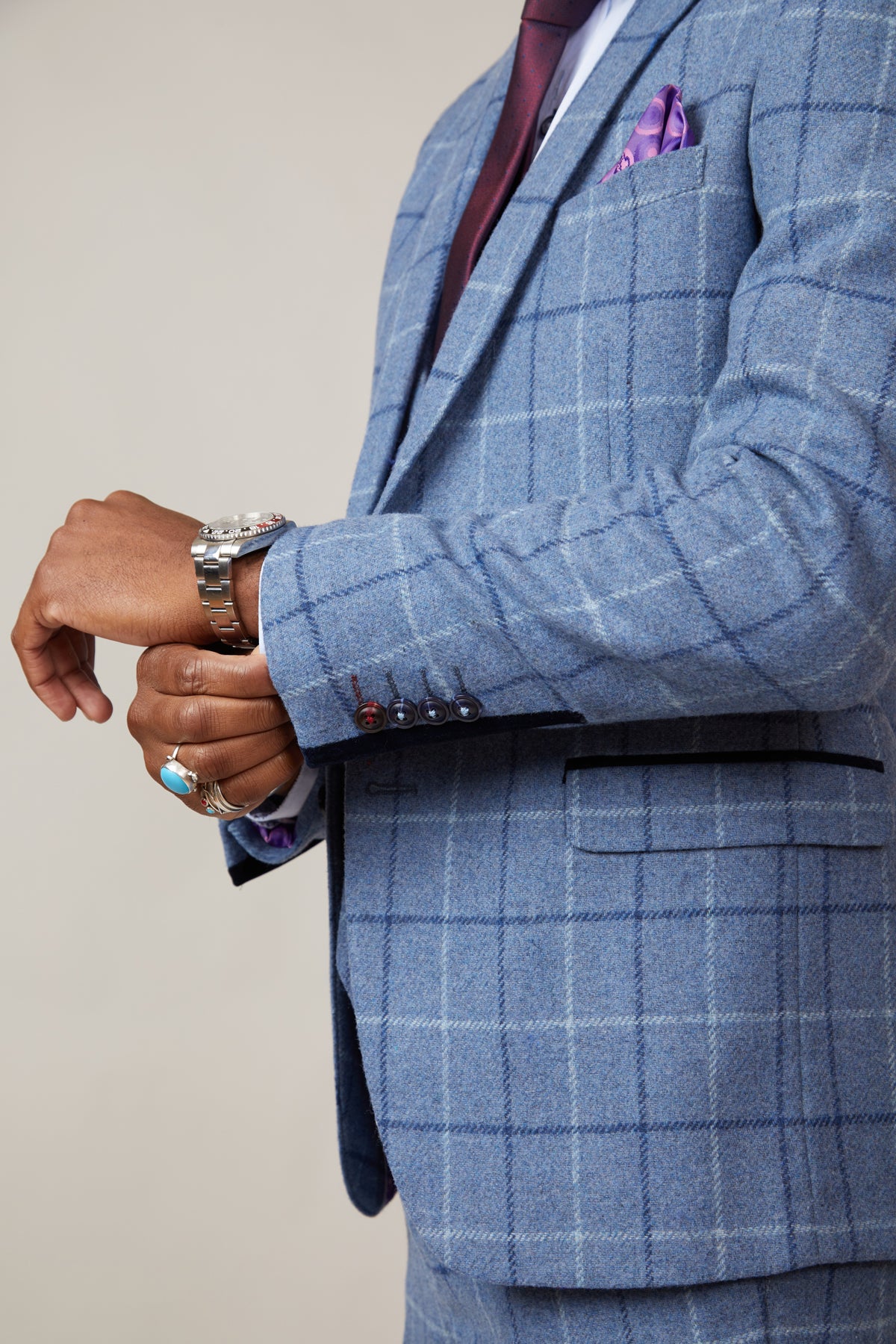 Wearing the marcdarcy CLINTON Blue Tweed Check Blazer, a person adjusts a silver wristwatch and sports a burgundy tie, purple pocket square, and turquoise ring against a softly blurred background for classic heritage style.