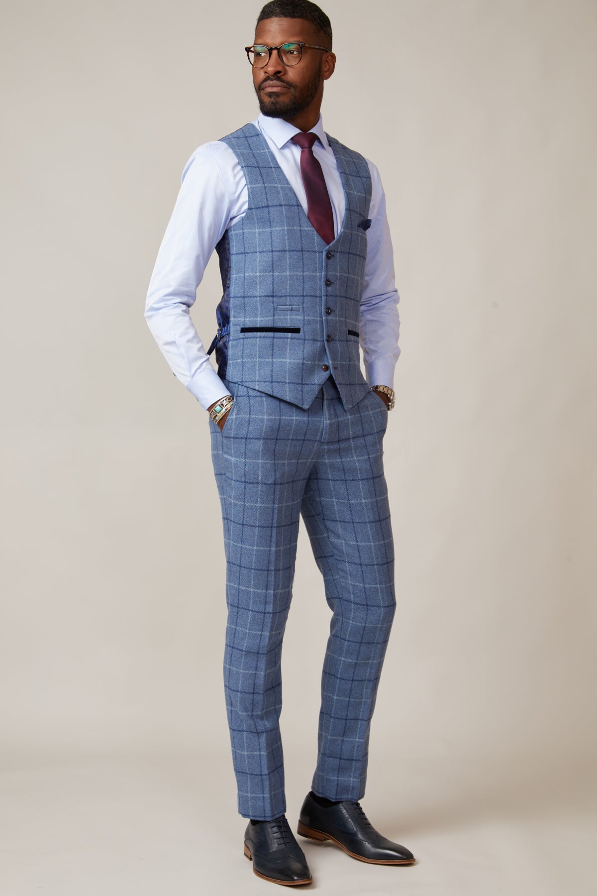 A man wearing glasses poses with his hands in his pockets, dressed in a blue check suit and marcdarcy's CLINTON Blue Tweed Check Waistcoat, white shirt, maroon tie, and black dress shoes against a plain background.