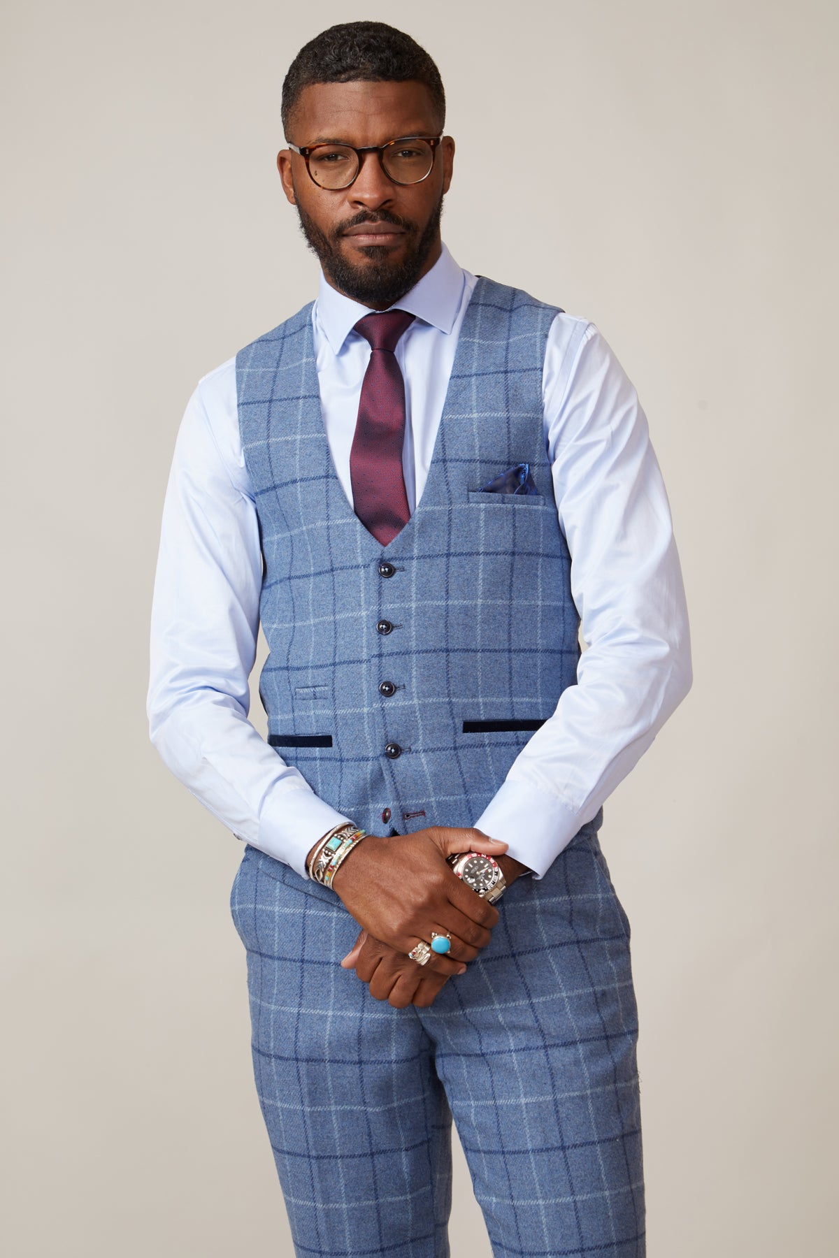 A man wearing the marcdarcy CLINTON Blue Tweed Check Waistcoat with matching pants, a white shirt, and a burgundy tie stands with his hands clasped. He accessorizes with glasses, bracelets, a watch, a ring, and sports a short beard and mustache.