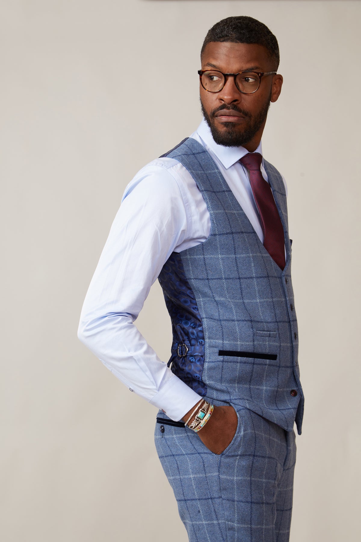 A Black man in a marcdarcy CLINTON Blue Tweed Check Waistcoat, white shirt, burgundy tie, with glasses and a bracelet.