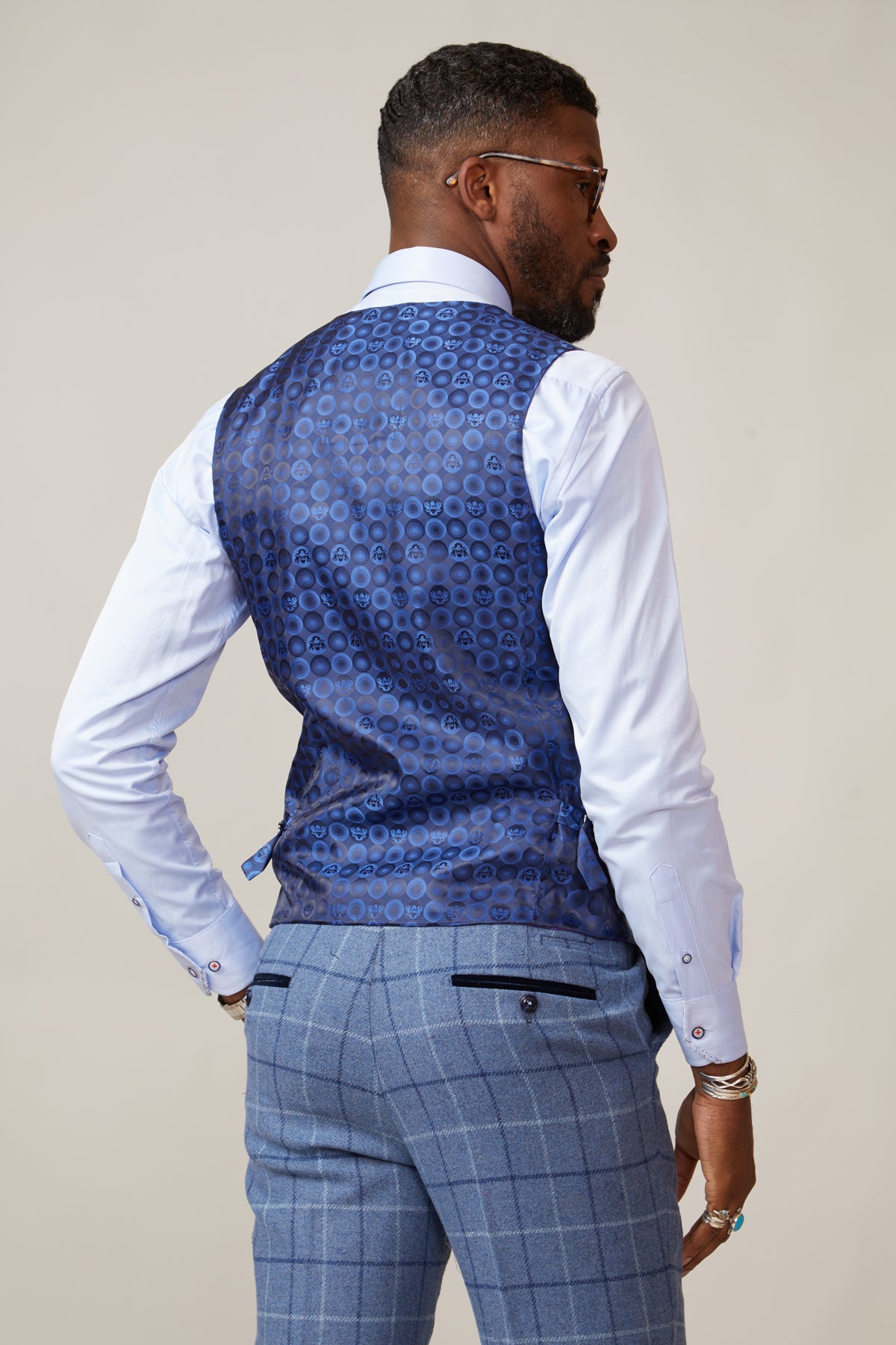 A man wears the marcdarcy CLINTON Blue Tweed Check Waistcoat with a white dress shirt and light blue checkered pants, facing away and turning his head slightly, accessorized with bracelets and sunglasses.