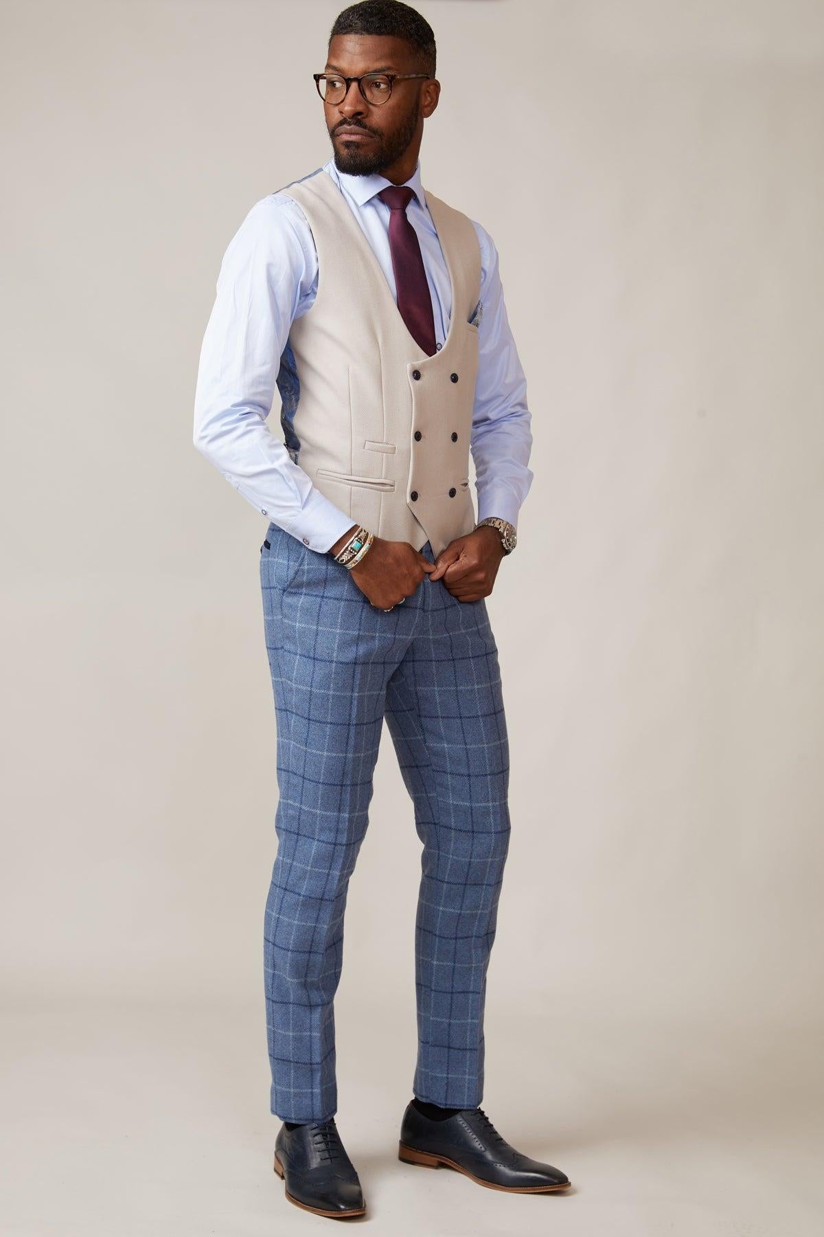 A man with glasses and a beard poses in a neutral studio wearing the marcdarcy KELVIN - Stone Double Breasted Waistcoat, a light blue shirt, burgundy tie, blue plaid pants, and black shoes, with one hand in his pocket.