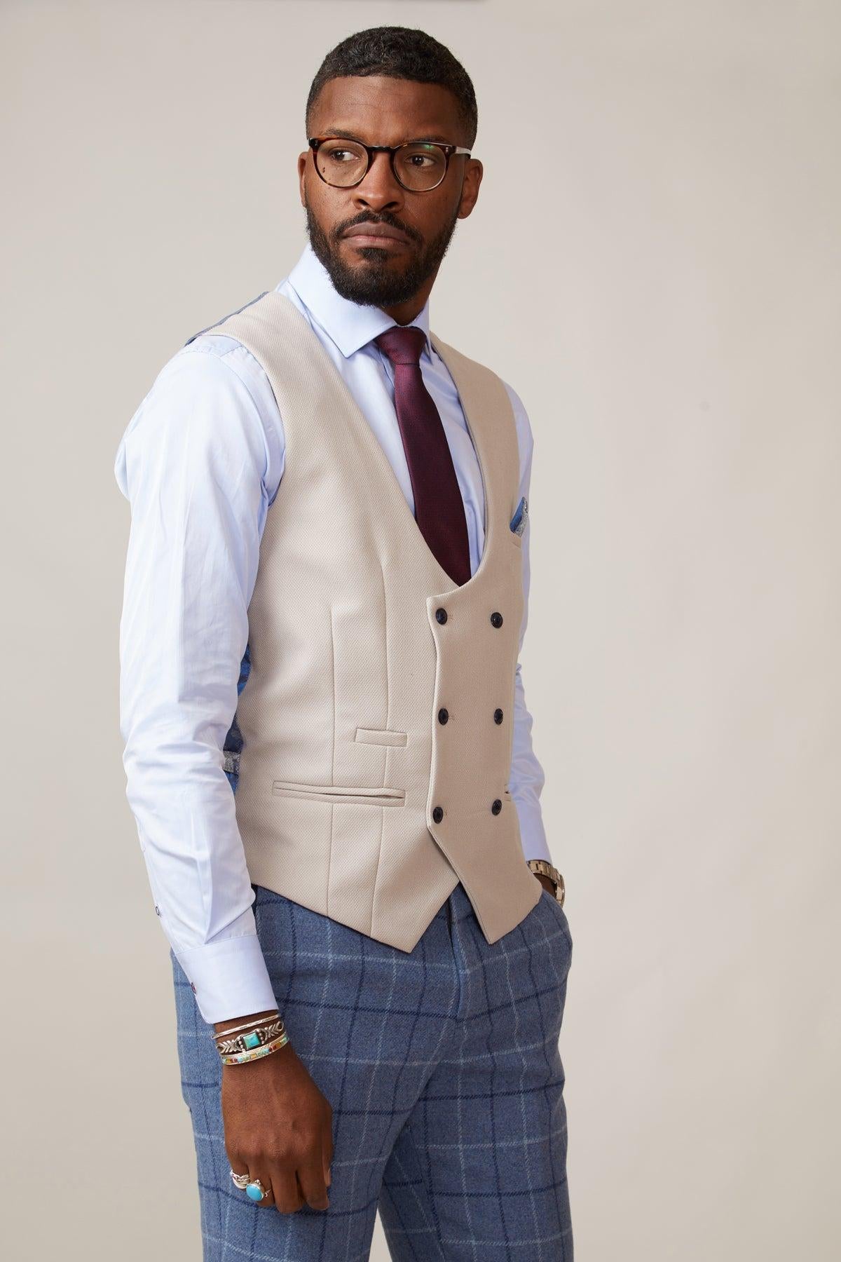 A man wears a marcdarcy KELVIN Stone Double Breasted Waistcoat, a light blue dress shirt, and slim fit blue plaid trousers, standing against a plain light background and looking to the side.