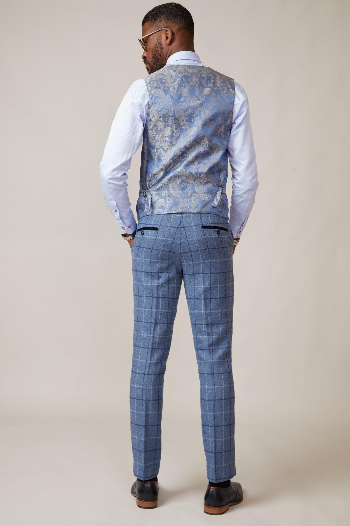 A man stands facing away, wearing the marcdarcy KELVIN Stone Double Breasted Waistcoat over a white dress shirt and blue plaid dress pants with dark shoes, set against a plain beige background.