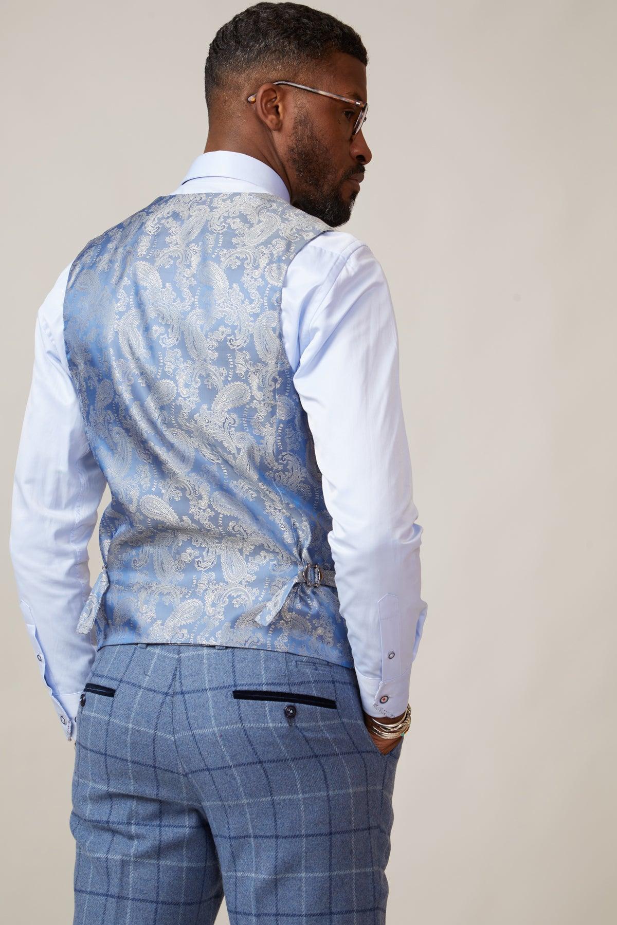A man faces away, wearing a slim fit, stone-colored marcdarcy KELVIN double breasted waistcoat over a white dress shirt and blue plaid trousers. He has glasses, neatly styled hair, and a beard against a plain light background.