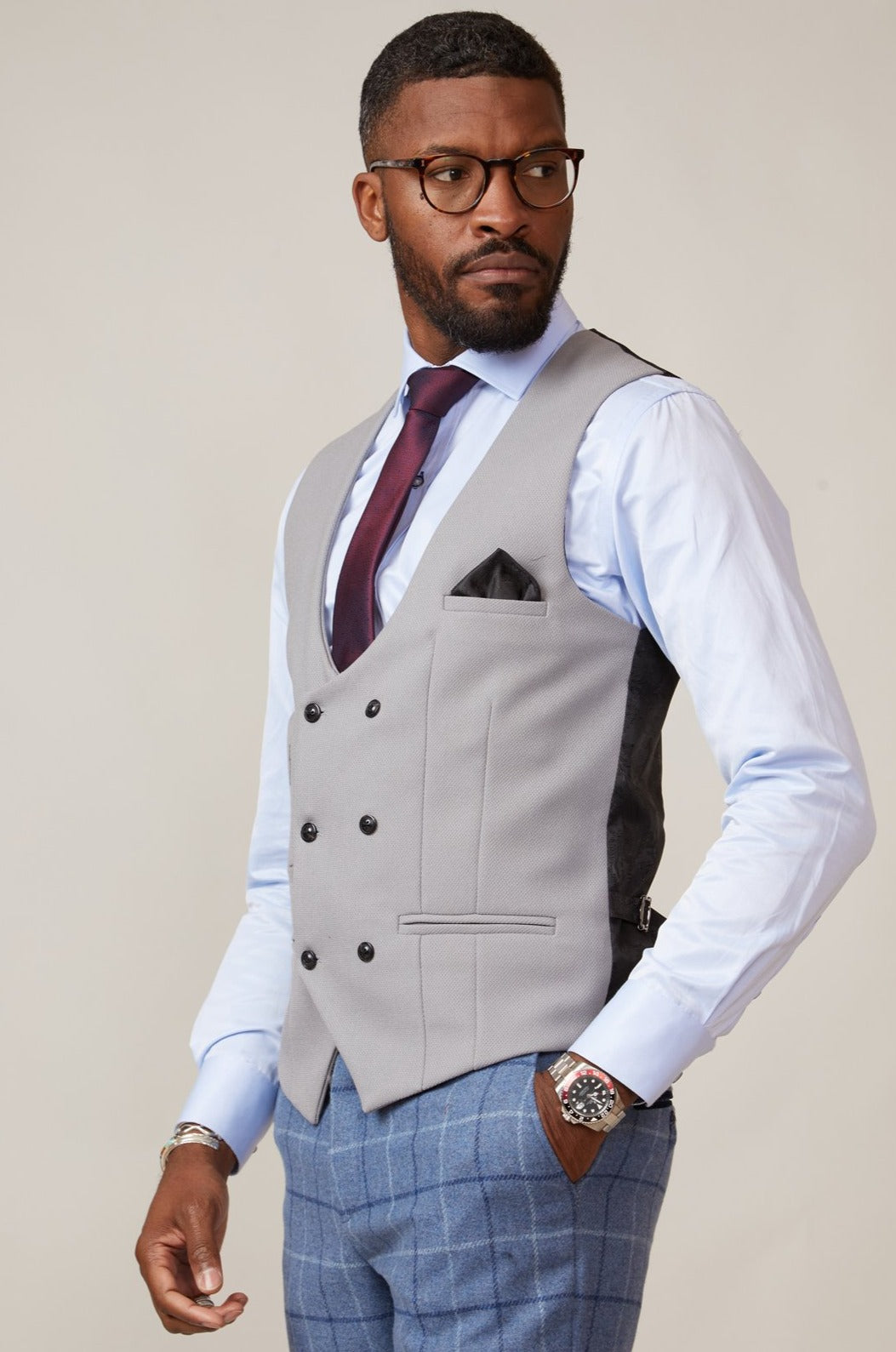 A man in glasses, a light blue shirt, the marcdarcy KELVIN Silver Double Breasted Waistcoat, burgundy tie, blue checkered pants, and a wristwatch stands turned slightly to the side, looking away from the camera.