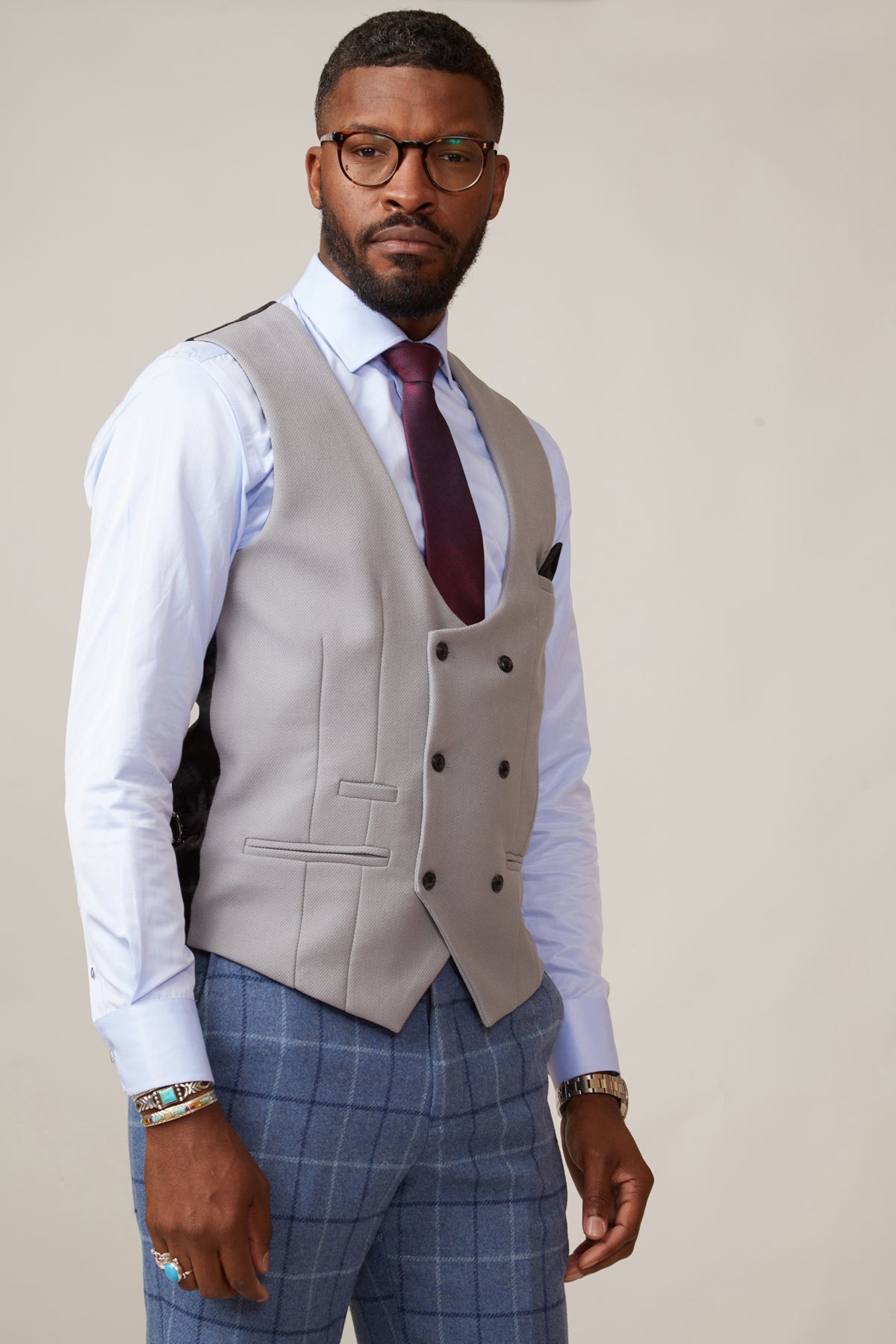 A man in glasses wears the marcdarcy KELVIN Silver Double Breasted Waistcoat, paired with a white dress shirt, burgundy tie, and blue plaid trousers, standing confidently against a plain light background.