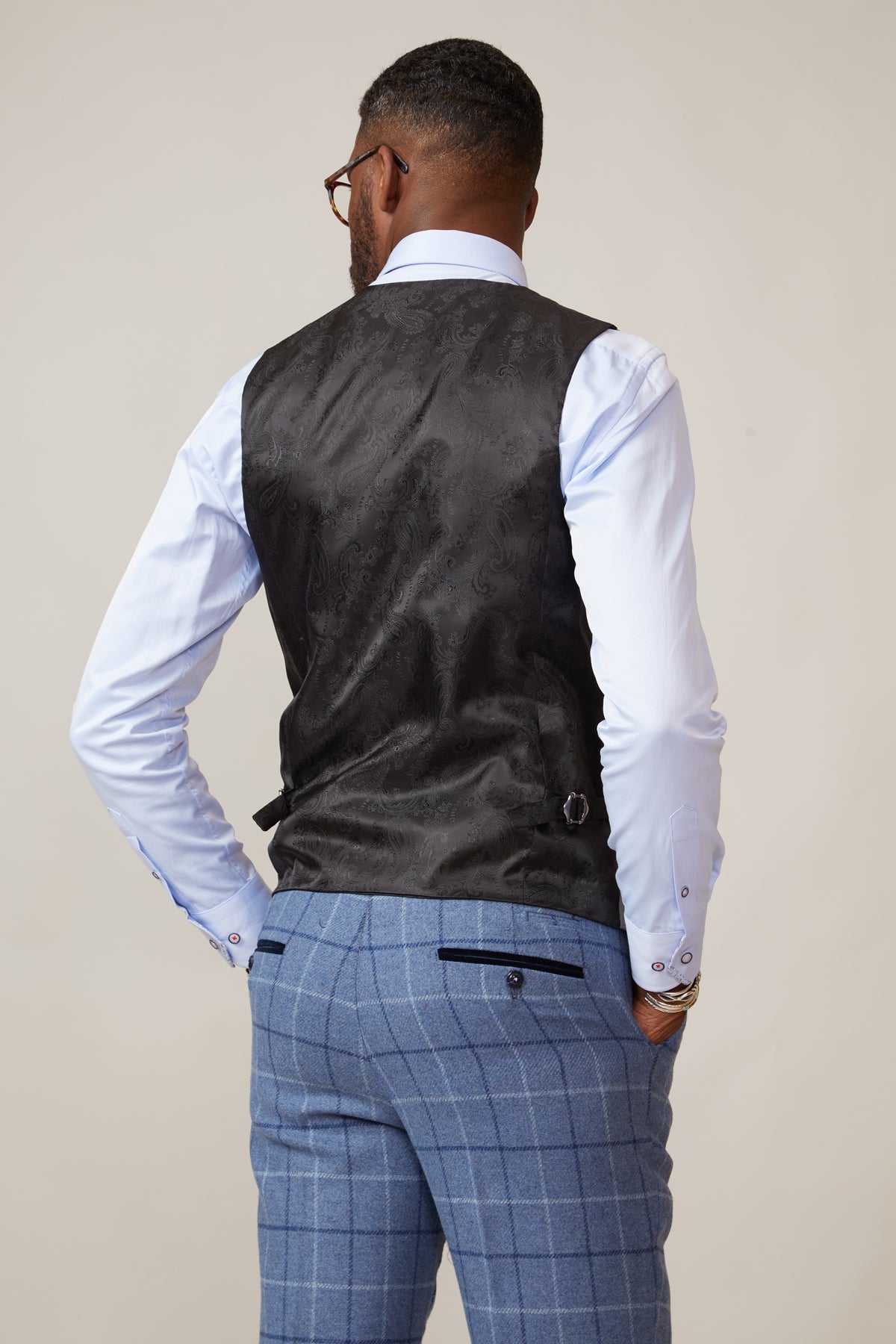 A man stands facing away, wearing the marcdarcy KELVIN Silver Double Breasted Waistcoat over a white dress shirt and light blue plaid trousers, his hands on his hips.