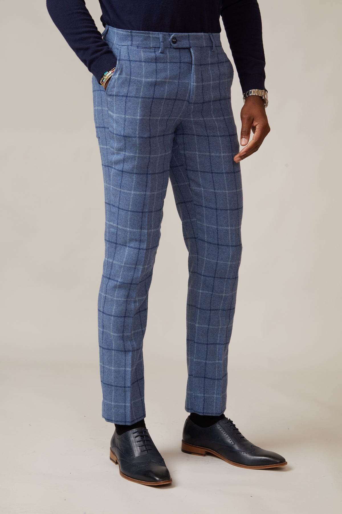 A person wearing marcdarcy's CLINTON Blue Tweed Check Trousers, paired with a dark long-sleeve shirt, black dress shoes, and bracelets, stands hands-in-pockets against a plain light background.