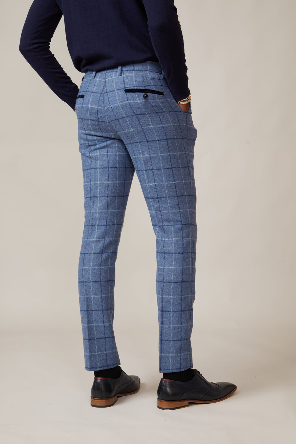A person stands with their back to the camera, wearing marcdarcy CLINTON Blue Tweed Check Trousers, a navy long-sleeve top, and black dress shoes against a light neutral background.