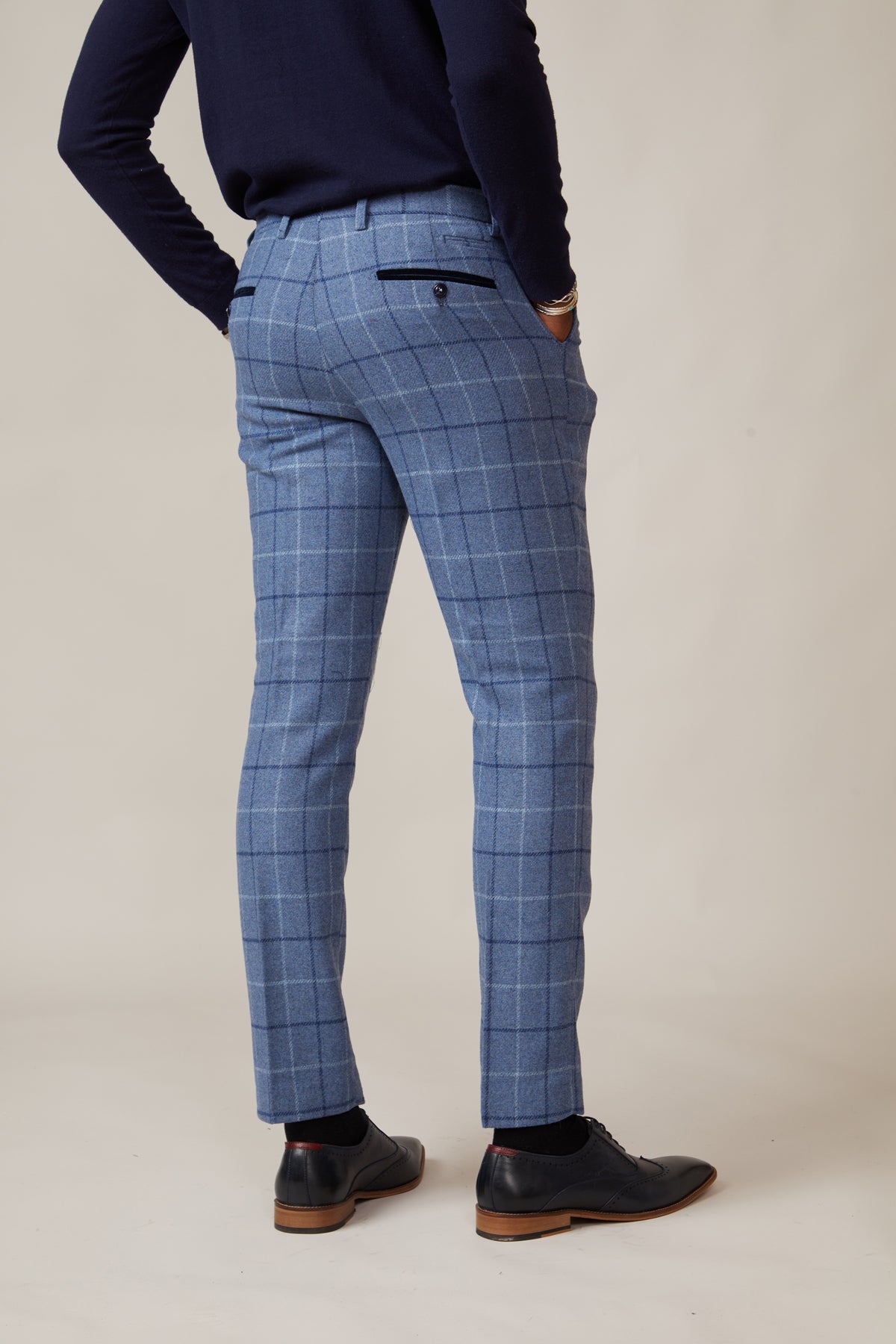 A person stands with their back to the camera, wearing marcdarcy CLINTON Blue Tweed Check Trousers, a navy long-sleeve top, and black dress shoes against a light neutral background.