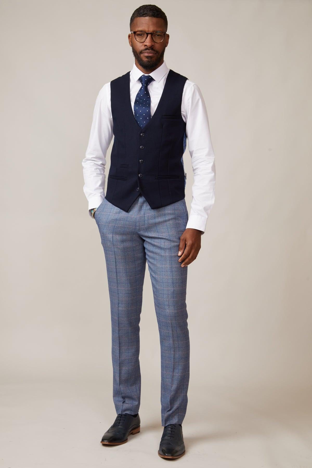 A man in glasses stands against a plain background wearing a slim fit white shirt, the marcdarcy KELVIN navy single breasted waistcoat, blue patterned tie, light blue plaid trousers, and dark dress shoes with his hands by his sides.