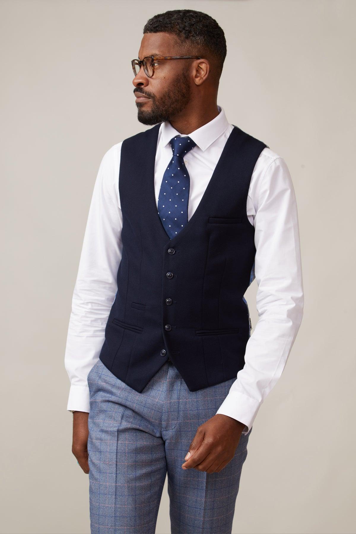 A man wearing glasses, the marcdarcy KELVIN Navy Single Breasted Waistcoat, a white dress shirt, blue polka dot tie, and gray plaid trousers stands against a plain light background, looking to his left.