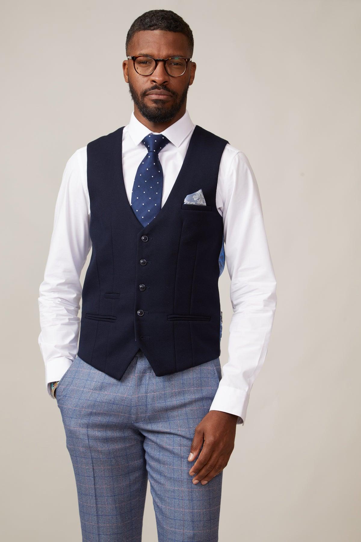 A man wears a slim fit white dress shirt, marcdarcy KELVIN navy single breasted waistcoat, blue polka dot tie, pocket square, and gray plaid trousers against a light background, confidently showcasing polished formalwear.