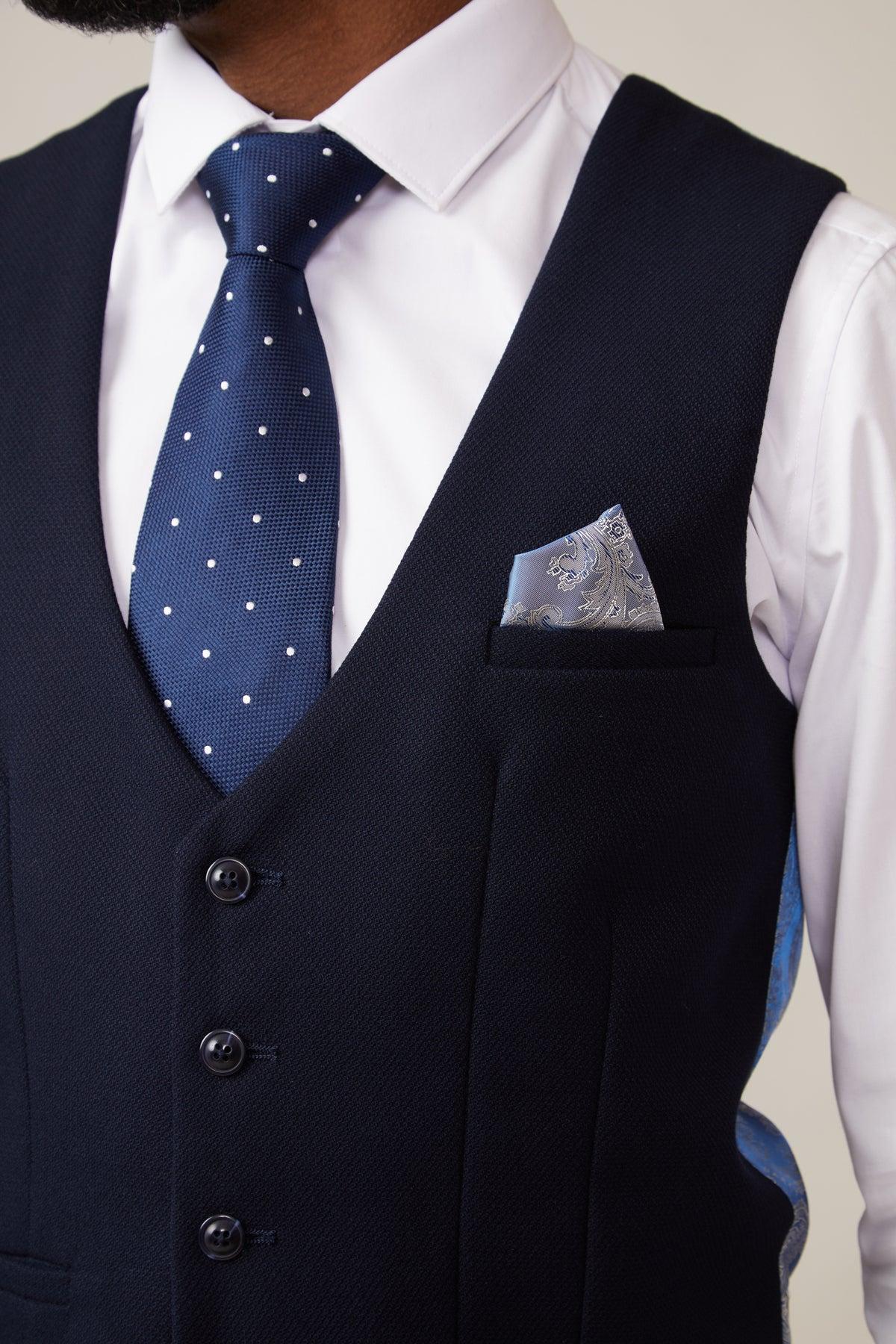 A person wears the marcdarcy KELVIN Navy Single Breasted Waistcoat over a white shirt, blue polka dot tie, and blue paisley pocket square—elegant formalwear. The persons face is partially out of frame.