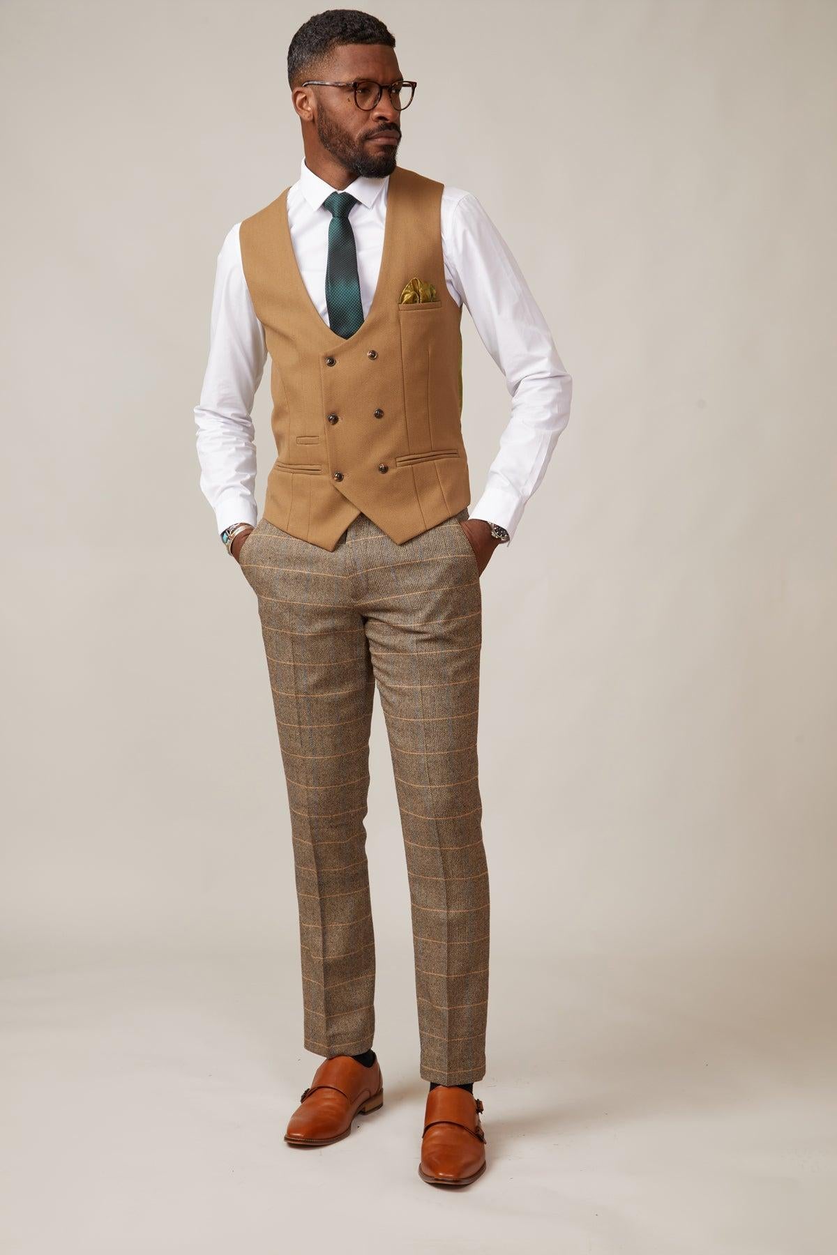 A man stands with hands in pockets, wearing a marcdarcy KELVIN - Oak Double Breasted Waistcoat with paisley lining, white shirt, dark green tie, plaid trousers, brown shoes, glasses, and a yellow pocket square.