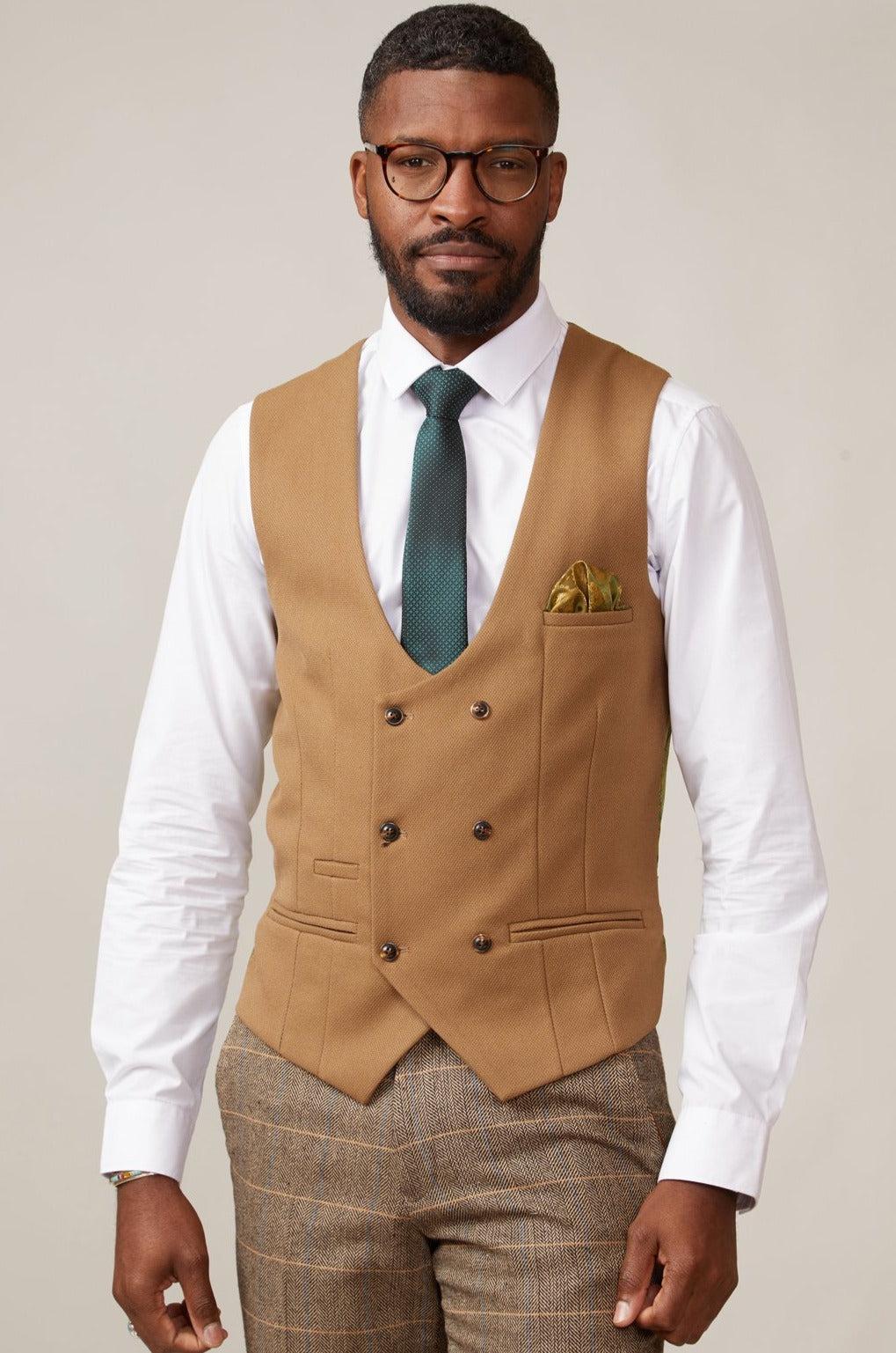 A man wearing glasses, a white dress shirt, green tie, KELVIN - Oak Double Breasted Waistcoat by marcdarcy with brown buttons, plaid brown trousers, and a yellow pocket square stands against a plain background.