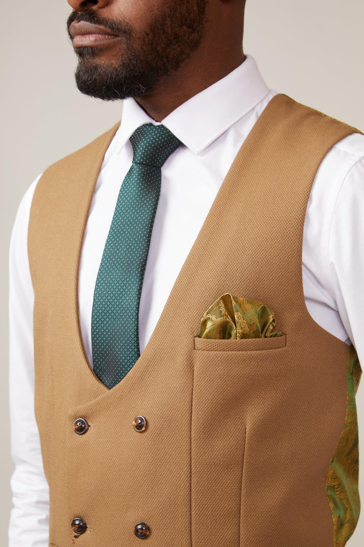 A man wears the marcdarcy KELVIN - Oak Double Breasted Waistcoat over a white shirt, styled with a green patterned tie and green-gold pocket square. The waistcoat features a subtle paisley print lining. Shown from shoulders to mid-chest.