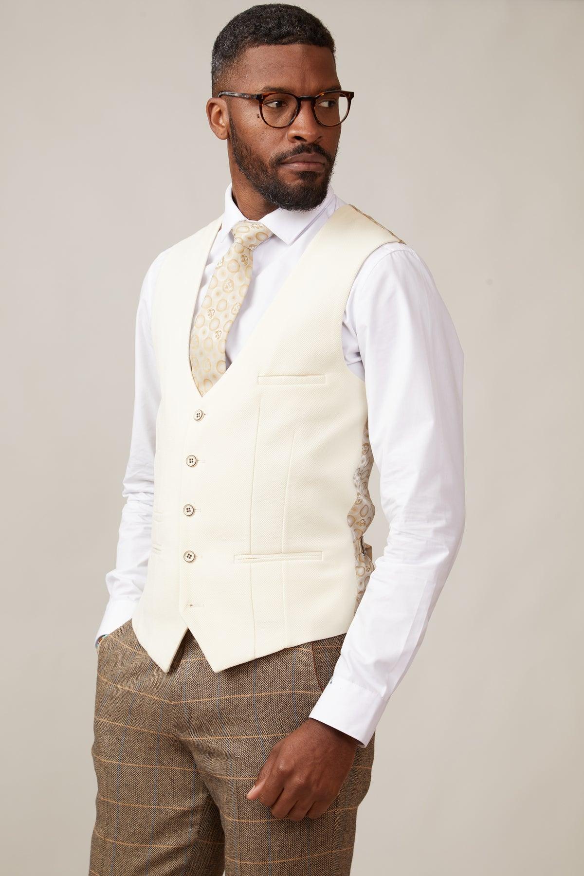 A man with short hair, beard, and glasses wears the marcdarcy TED Tan Tweed Check Suit with Kelvin Cream Waistcoat—styled with a white shirt and beige tie—standing against a plain background, hand in pocket.