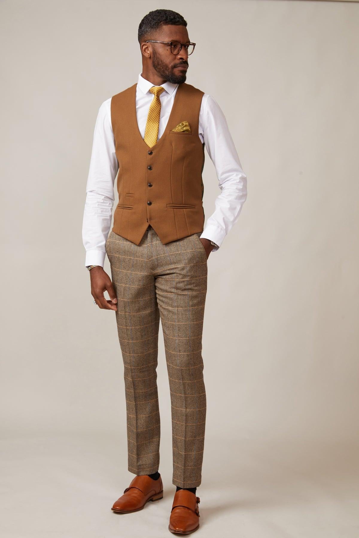 A man wearing the marcdarcy KELVIN Tan Single Breasted Waistcoat, a white shirt, yellow tie, plaid brown trousers, and brown dress shoes stands against a beige background, looking to his left.