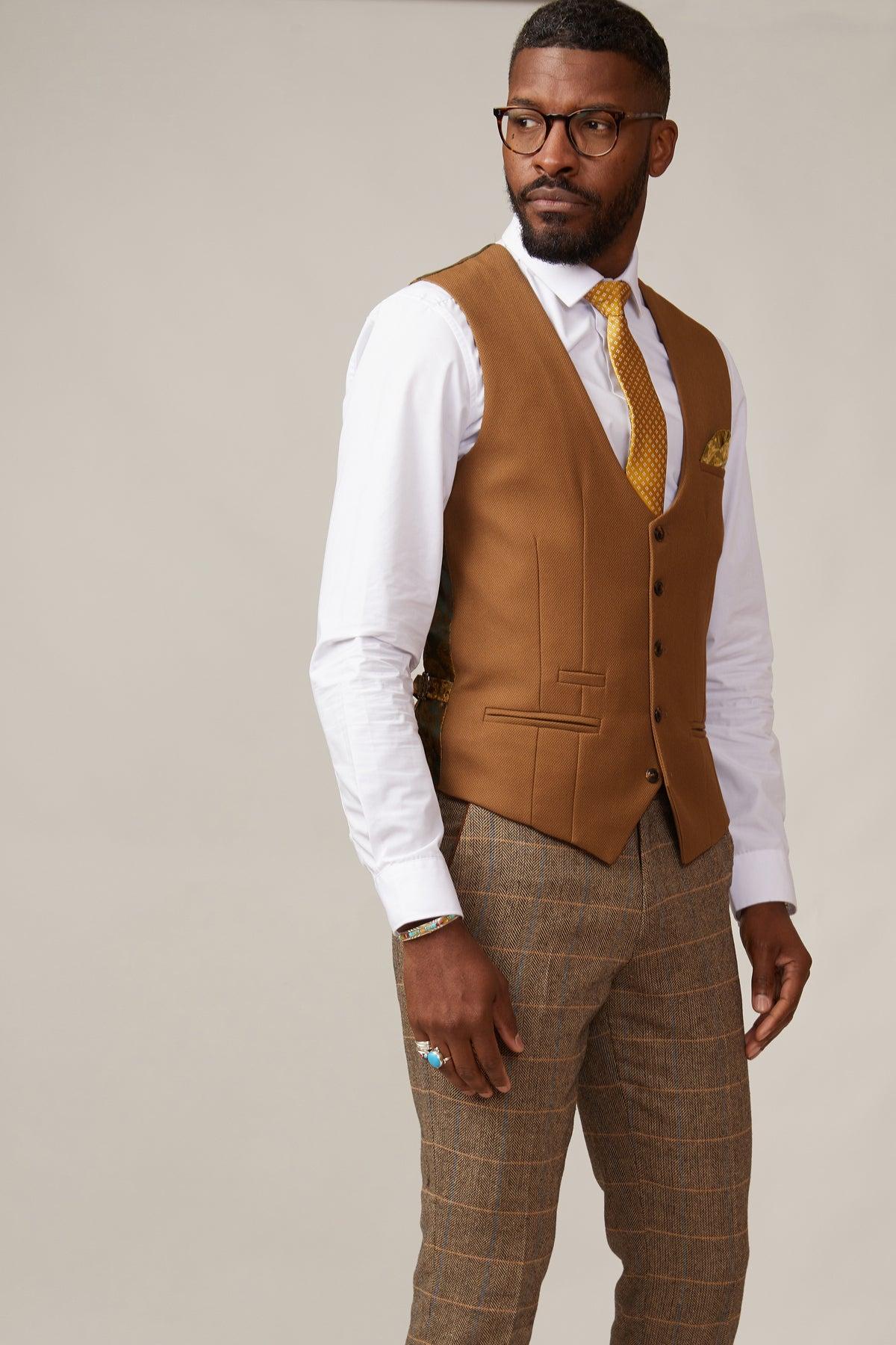 A man wearing the marcdarcy KELVIN Tan Single Breasted Waistcoat, glasses, a white shirt, mustard tie, and plaid brown trousers stands against a neutral background, looking to the side.