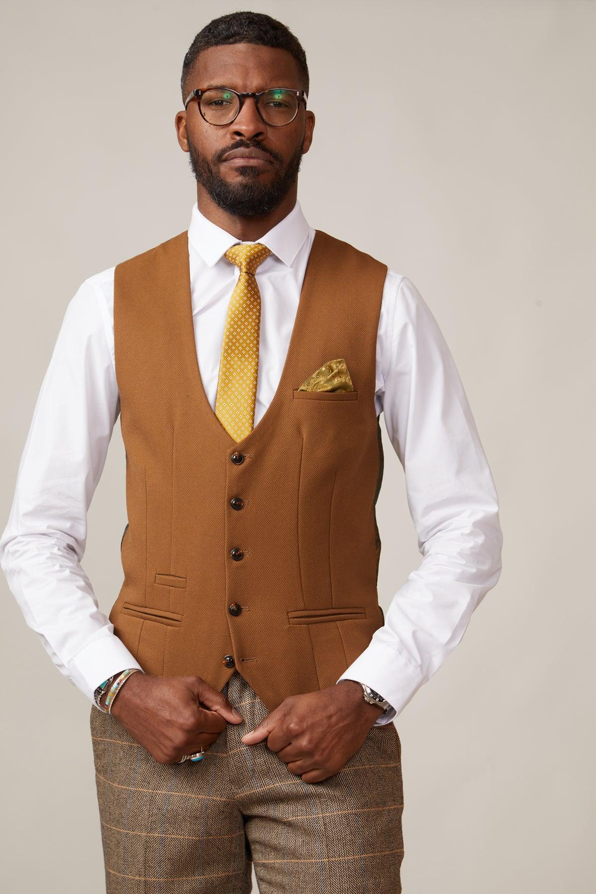 A man in glasses wears a slim fit white dress shirt, brown plaid pants, and the marcdarcy KELVIN tan single breasted waistcoat with buttons, paired with a yellow patterned tie and matching pocket square, against a plain light background.