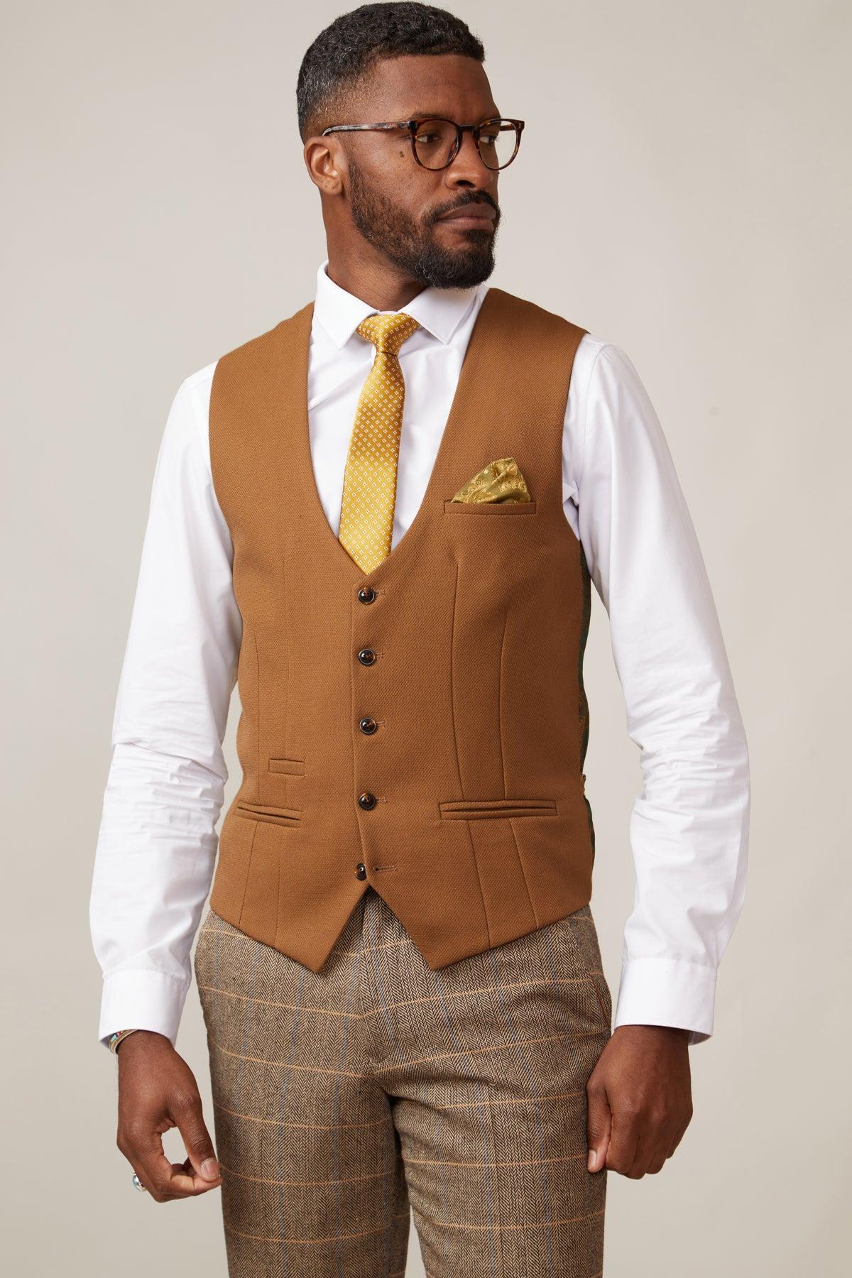 A man wearing glasses, a white shirt, the marcdarcy KELVIN Tan Single Breasted Waistcoat, a yellow patterned tie with matching pocket square, and brown plaid trousers stands against a plain background.
