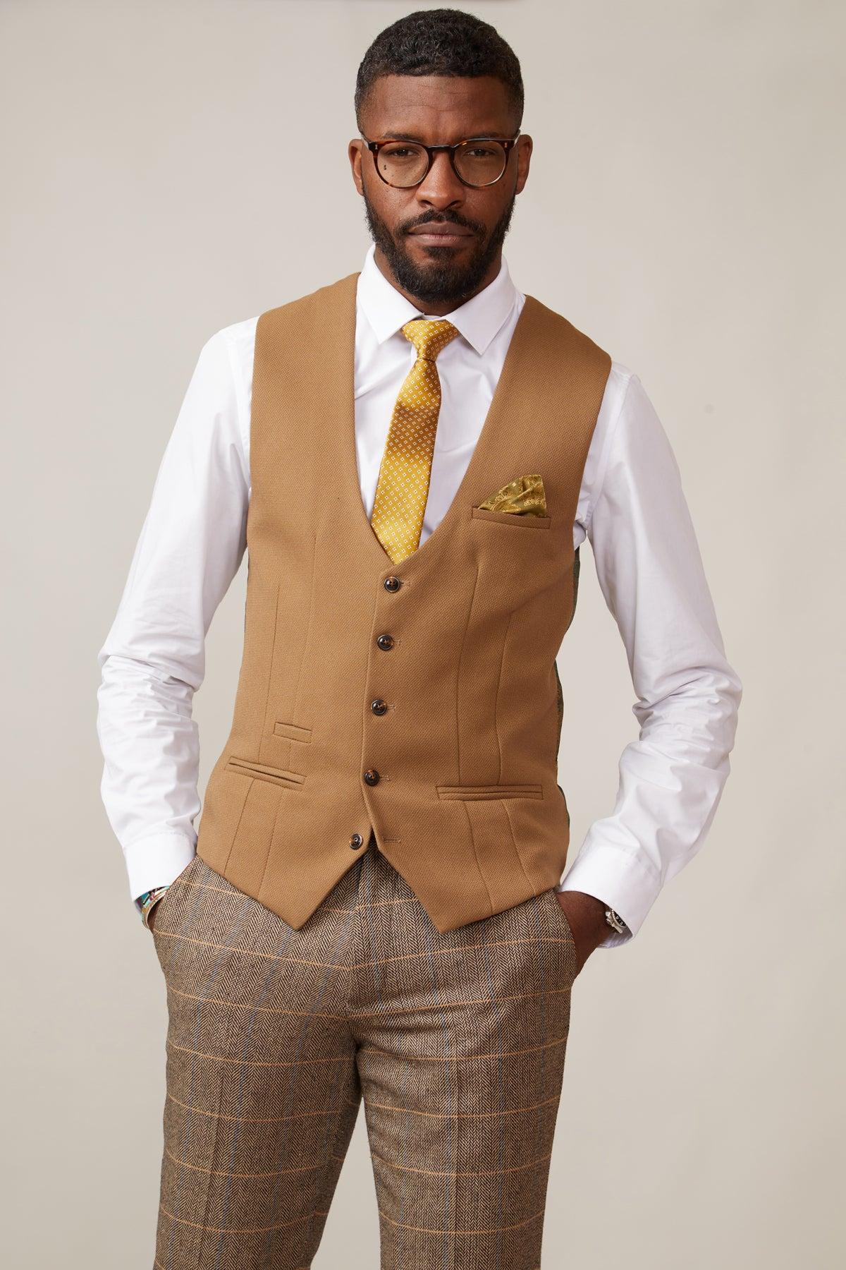 A man with short hair and a beard, wearing glasses, a white shirt, yellow patterned tie, and brown plaid pants, showcases the marcdarcy KELVIN - Oak Single Breasted Waistcoat with matching pocket square against a plain background.