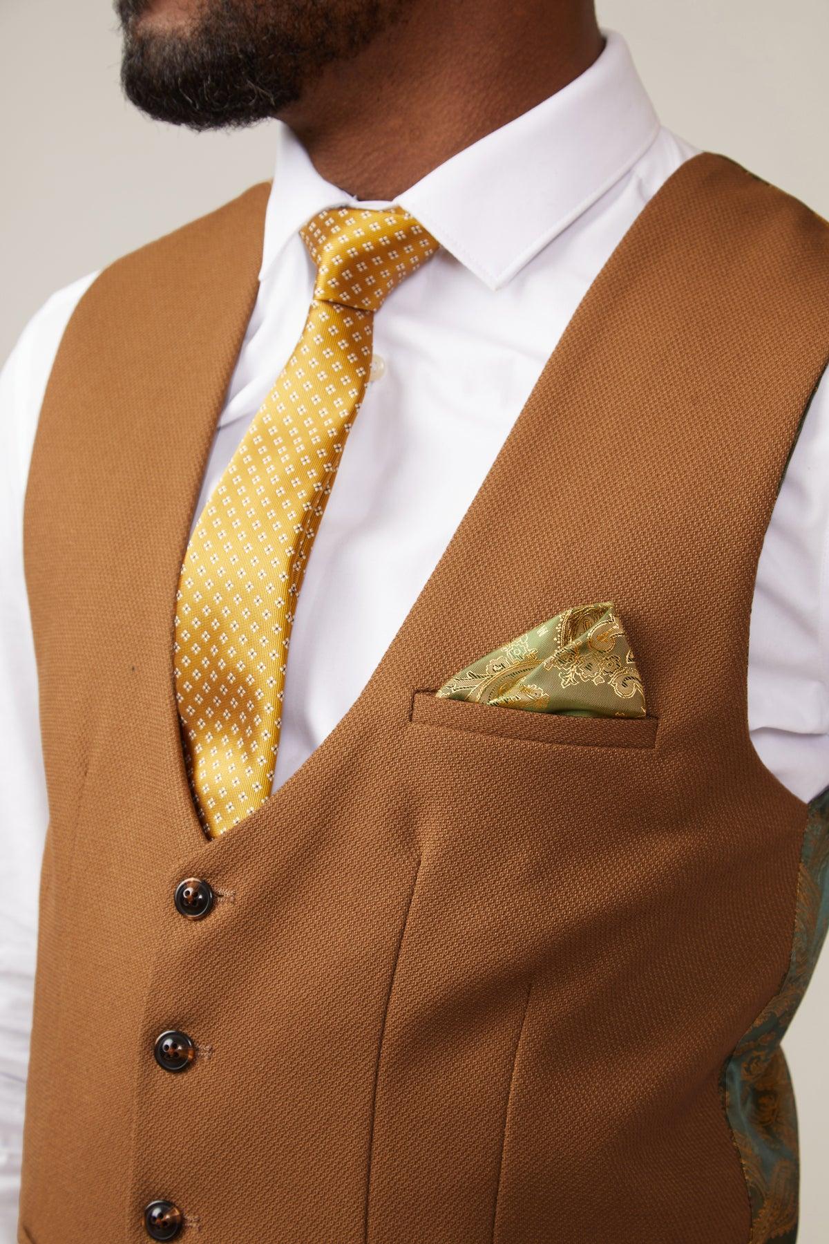 A man wears the marcdarcy KELVIN Tan Single Breasted Waistcoat over a white shirt, styled with a yellow patterned tie and green pocket square. Only his lower face and torso show. The waistcoat has an elegant paisley print lining.
