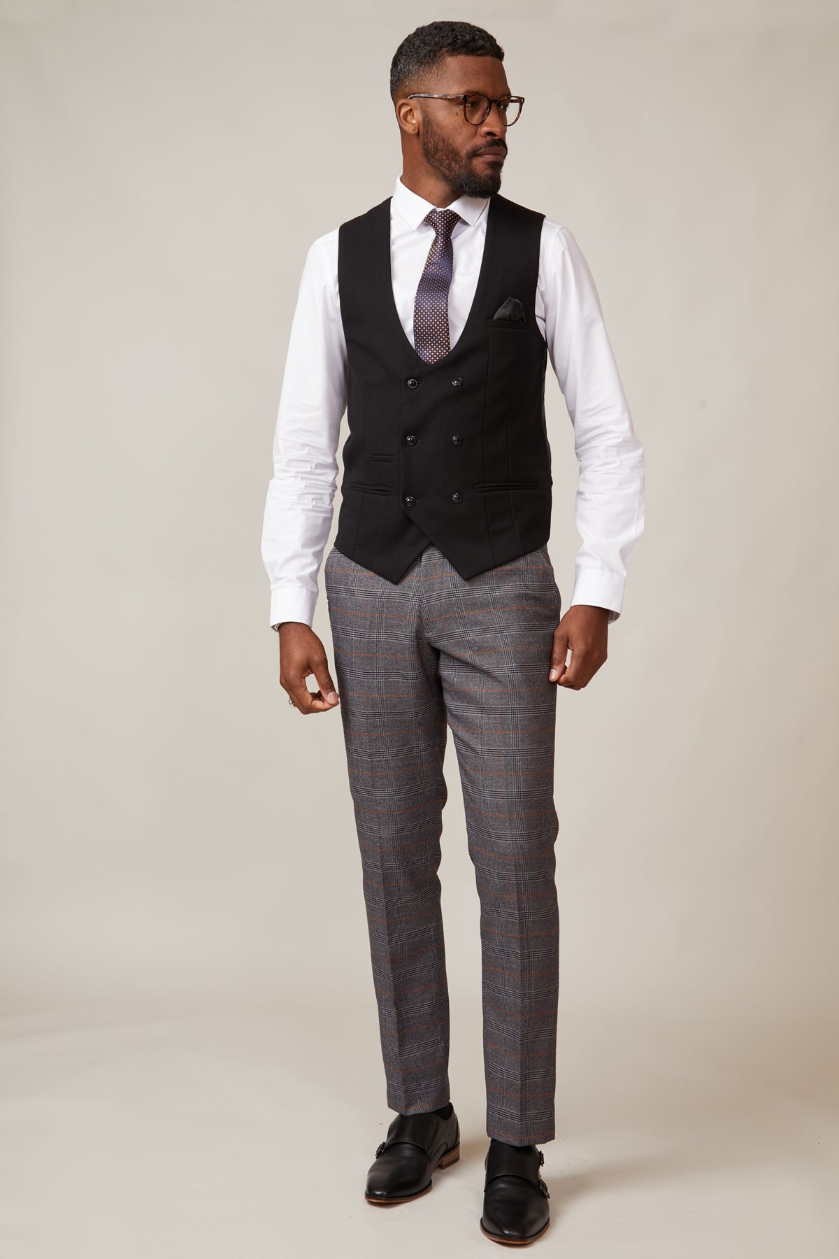 A man in glasses stands against a plain background, wearing a white shirt, patterned tie, gray plaid trousers, black dress shoes, and the marcdarcy KELVIN Black Double Breasted Waistcoat. He looks to the side with one hand in his pocket.